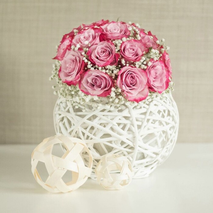 This image presents a serene, contemporary floral display that could easily belong on a carefully styled shelf in a Grove Park home, with a lush bouquet of pink roses and white baby's breath cradled in a distinctive white woven vase. At the centre of the scene is a rounded cluster of roses, each bloom generously open, revealing layers of overlapping petals. The colour shifts gently from a delicate, almost porcelain blush at the inner petals to a more intense, raspberry-pink edge, creating a natural two-tone effect. Interspersed among the roses are airy sprays of white Gypsophila, their tiny blooms sprinkled throughout like soft, floral confetti, giving the bouquet a cloud-like lightness and a classic, romantic feel. The flowers are arranged in a spherical white container woven from slender strips that resemble rattan or wicker painted bright white, with open spaces between the strands. This lattice design allows the light to dance through, throwing subtle patterns on the smooth, white surface beneath, suggesting a polished tabletop or console. To the left and slightly forward stand two decorative spheres in pale beige, crafted from thin, interlaced natural fibres. One is notably larger, the other smaller, together echoing the organic textures of the vase in a quieter, neutral palette. Behind the arrangement, a warm, cream-beige wall with a gentle texture serves as an unobtrusive backdrop, helping the pink and white tones stand out without any visual noise. The lighting is soft and even, as if filtered through a nearby window, lending the scene a calm, welcoming glow. It feels like a bouquet you might order from a trusted local florist for same-day delivery across Grove Park to celebrate a birthday, a quiet anniversary dinner, or to brighten a new flat with understated romance.