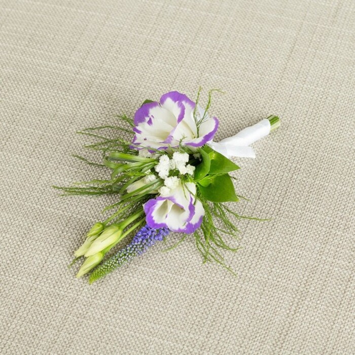 A single, artfully crafted boutonnière is laid on a softly textured light beige fabric, the weave of the linen-like background providing a gentle contrast to the smooth petals and adding a warm, neutral tone to the scene. At the centre, two lisianthus flowers open fully, their creamy white petals elegantly ruffled and traced with a distinct margin of deep purple, like delicate brushstrokes that follow every fold and curve. Tucked between and around them are fine clusters of tiny white filler blooms reminiscent of baby's breath, giving the arrangement an airy halo that lightens the overall look. Standing upright along one side, a narrow spike of muscari introduces a line of tiny, grape-like bells in a rich violet-purple, adding both height and a slightly wild, garden-inspired character. Surrounding these flowers, fine-textured greenery-likely asparagus fern-spills softly outward, its feathery fronds mingling with slightly broader green leaves to create layers of fresh, natural foliage. Two slender, pale green buds rest at an angle, not yet opened, quietly symbolising new chapters and the unfolding of the day's events. The exposed stems gather neatly at the base, where they are tightly wrapped in crisp white satin ribbon, smooth and luminous against the greens and purples, and secured with a modest, well-tied bow that makes it ready to pin on a lapel. The mood is one of thoughtful elegance and calm celebration, perfectly suited to a groom or guest attending a wedding in Grove Park, perhaps walking from a nearby townhouse to a local venue. The careful balance of white, purple, and green reflects the skill of a local florist who understands how to create something that feels both timeless and uniquely personal.