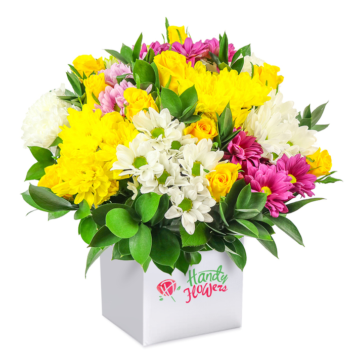 A full, rounded bouquet of mixed flowers overflows from a modern white cube box, presented against a pure white background that accentuates its bright and harmonious palette. The overall shape is a generous dome, tightly packed so that only glimpses of the stems and container are visible beneath the profusion of petals. The most striking feature is the abundance of yellow: large, multi-layered chrysanthemums and clusters of small yellow spray roses bring warmth and optimism, their petals soft and cushiony, some still in bud, suggesting freshly trimmed stems. Nestled among these are cool white flowers-delicate daisy-like blooms with distinctive green centres and similarly sized white chrysanthemums with soft, feathery petals-that provide clean contrast and give the design an airy, open feel. Threaded artfully throughout, deep fuchsia and magenta chrysanthemums, occasionally brushed with lighter pink tips, add pockets of jewel-like intensity and lend a contemporary twist to the colour story. Supporting this colourful canopy is a ring of dark green foliage, likely ruscus or another sturdy greenery, its glossy leaves arching outward and upward, framing the flowers and adding a sense of depth and structure. The container itself is a smooth, cube-shaped white gift box whose simplicity keeps the focus firmly on the blooms; its front panel bears a neat floral emblem in red alongside flowing script in green and red, suggesting a carefully branded creation from a local florist. The image evokes a sense of cheerful generosity, the kind of uplifting arrangement someone in Grove Park might have delivered to a friend near the river, a home off Grove Park Terrace, or a gathering at a nearby family table, marking celebrations, get-well wishes, or simple acts of kindness.