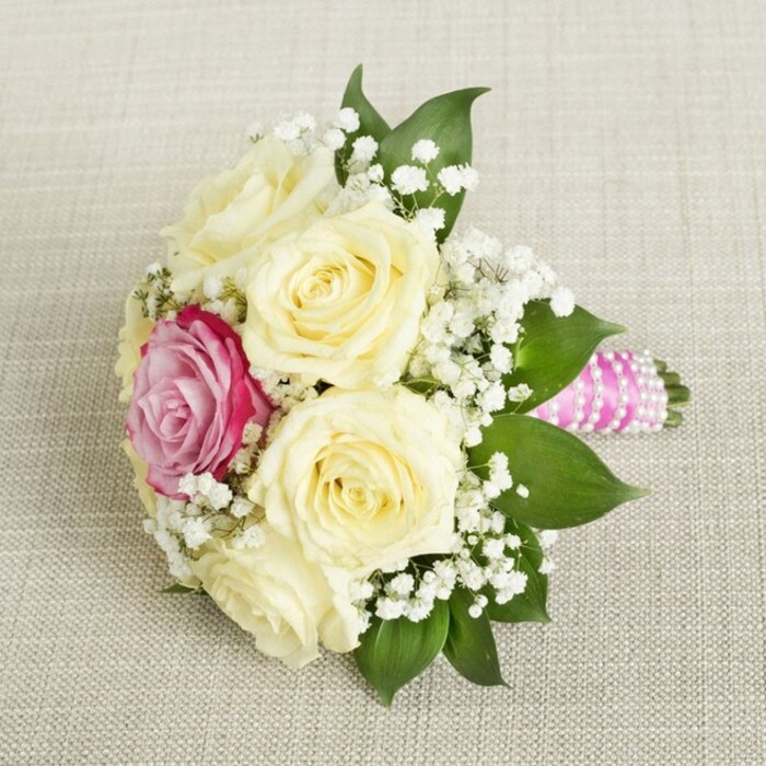 Viewed from above and gently angled, a beautifully tailored bouquet is laid atop a muted linen cloth, the type often seen at celebrations near Grove Park's leafy avenues. At its heart are five or six creamy, pale yellow roses, their petals just starting to unfurl - testament to their freshness and skilled local sourcing. A single, radiant fuchsia-pink rose stands out just to one side, commanding attention with its vibrant colour, much like the seasonal blossoms gracing Dukes Meadows in springtime. Liberally interspersed with airy clusters of white baby's breath, the bouquet radiates lightness and romance, complemented by the rich undertones of glossy dark green leaves circling the blooms. The bouquet's handle is expertly wrapped in bright pink satin ribbon, further accentuated by dainty white pearls spiralled with care - a finishing touch inspired by Grove Park's classic-meets-contemporary style. Resting on the soft, neutral fabric, the vibrant arrangement invites admiration for its balance and warmth, ideal for weddings, bridal parties, or as a feature in articles about floral design, seasonal colour palettes, or local celebrations along Chiswick High Road.