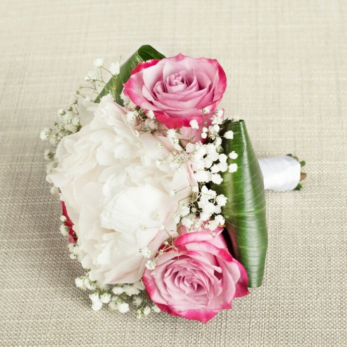 Captured from an artful, slightly elevated angle on a woven beige backdrop, this standout bouquet is much like something you'd find at a springtime wedding in Grove Park-timeless, elegant, and delicately composed. The centrepiece: one plush, white peony brimming with creamy, ruffled petals-its abundance radiates a softness akin to early morning clouds drifting over the local green. Flanking the peony, two open pink roses offer bold contrast, their petals marked by a dramatic gradient from rich fuchsia edges fading beautifully into notes of pale blush and even whispers of lavender, a true highlight of colour and freshness. Wisps of gypsophila, or baby's breath, serve as both filler and feature, their airy white blossoms tucked among the larger blooms to soften the composition and create a fairy-tale lightness. A graceful collar of deep emerald leaves-folded and glossy-encloses the arrangement, providing structure and vibrancy reminiscent of Grove Park's riverside foliage. The handle, bound in white satin, is neat and discreet, alluding to the skill and care of local florists who value both form and function. Bathed in natural, even light, every petal and curve is clearly defined, making this bouquet an inviting choice for weddings, milestone celebrations, or romantic gestures all across the Grove Park area.