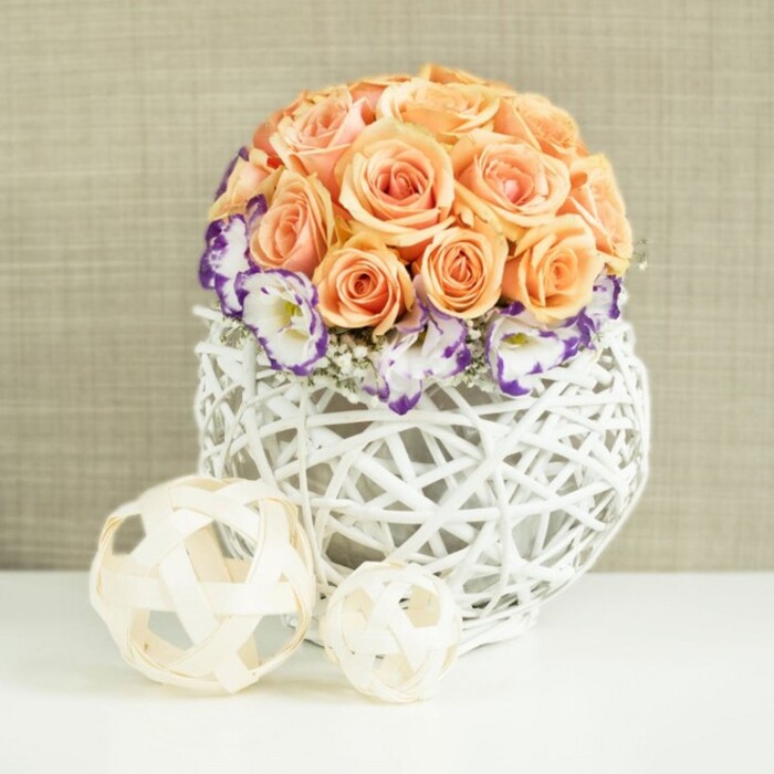 A beautifully curated floral centerpiece stands on a smooth white surface, its form and colours radiating a soft, celebratory calm that would suit a small wedding reception in Grove Park or an elegant dinner near Chiswick. At its core is a densely packed, spherical dome of roses in gentle peach and apricot shades, each bloom open and full, with layers of petals unfurling in soft curls. The roses are arranged so closely that they create a unified, rounded silhouette, like a single, luxurious rose cushion. Framing the lower edge of this dome is a collar of lisianthus in creamy white, each petal tipped with a vibrant border of violet and purple, adding intrigue and contrast without overwhelming the peach tones above. Fine sprigs of white gypsophila are nestled among the lisianthus, their small, cloud-like flowers adding lightness and a whisper of movement. The flowers emerge from a striking white woven vase with a wide, rounded belly, constructed from interlaced wicker or rattan strands that form an airy, open lattice, offering glimpses of stems and the shadows they cast within. On the white surface in front of the vase, two decorative spheres crafted from pale, natural-coloured strips of wood or rattan are placed side by side, one slightly larger than the other. Their open, crisscrossed pattern echoes the vase and gently leads the eye across the composition. Behind everything, a soft beige, slightly textured backdrop - reminiscent of linen or finely woven fabric - envelops the scene in warmth. The overall impression is one of quiet luxury and thoughtful design, ideal for wedding table arrangements or refined home décor throughout Grove Park, where understated elegance is often preferred over excess.