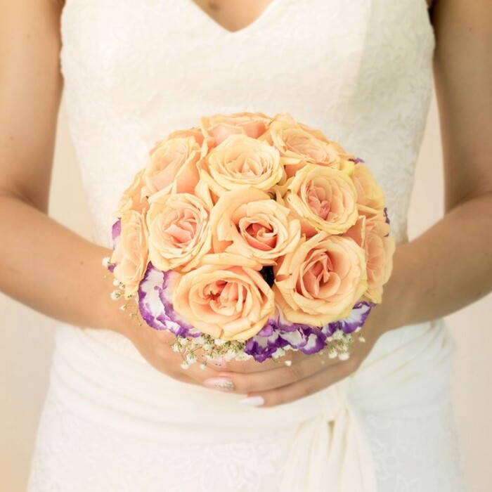 A detailed, close-up photograph focuses on the midsection of a bride, as though you are standing just in front of her in a quiet room somewhere in Grove Park, seeing only her wedding dress and bouquet. Her gown is a classic shade of white or ivory, with intricate lace overlaying the bodice and forming a soft V-shaped neckline that flatters without revealing too much. The lace pattern is delicate and feminine, a mix of small floral motifs and fine, net-like textures that sit smoothly against her skin. Around her waist, the fabric wraps and folds gracefully, creating gentle movement and visual interest before flowing down into a skirt of soft, textured layers that blur into the background. In her lightly clasped hands she holds a round bridal bouquet that seems to glow with warm, romantic colour. At the centre is a lush mass of roses in soft peach and apricot shades, some leaning toward blush pink. Their petals are tightly spiralled yet full, with a subtle gradient from pale outer edges to more intense, warm-toned cores, giving the impression of depth and richness. Underneath this dome of roses, a contrasting band of deep purple blossoms with creamy white centres-likely lisianthus-forms a ruffled collar. This purple-and-cream ring neatly frames the roses and introduces a cooler tone that makes the central flowers appear even more luminous. Around the base, tiny clusters of white filler flowers reminiscent of baby's breath peek out, adding an airy, lacy fringe that echoes the detailing on her dress. The bride's nails are softly manicured, with one thumb showing a glimmer of ombré sparkle, a small but personal touch. The light in the image is gentle and diffuse, revealing true colours and soft shadows, as if taken indoors near a window or in a calm corner of a Grove Park venue. The bouquet itself appears thoughtfully composed, evoking a sense of quiet joy and timeless romance suitable for wedding photography, local bridal features, or inspiration for couples planning their ceremony in and around Grove Park.