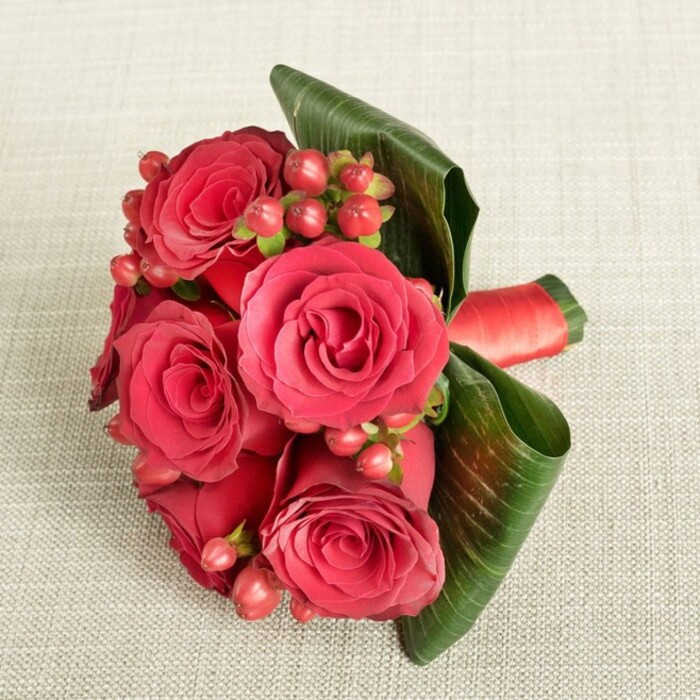 A richly romantic bouquet of deep red roses and berries lies on a pale beige fabric, whose fine, slightly raised texture resembles a soft cotton or linen throw often found in cosy Grove Park homes. The arrangement is compact yet full, with roughly six large roses gathered closely together, each one in glorious full bloom. The petals are thick and velvety, spiralling outward from tight, shadowed centres into more open, ruffled edges, showcasing an array of red tones from dark, wine-like crimson to brighter scarlet where the light grazes the tips. Tucked between the roses are generous clusters of small, rounded Hypericum berries, shading from deep red to subtle pinkish-red, attached to slender green stems that give a delicate, almost woodland texture. These berries introduce a cheerful, slightly playful note that lightens the drama of the roses. Encircling the base, two or more broad, dark green leaves are artfully curled and wrapped, their smooth surfaces and visible veins forming a strong, sculptural border that sets the flowers off with striking contrast. Around the stems, a neatly tied red satin or silk ribbon provides structure, its slight sheen catching the light and echoing the luxurious feel of the petals. The bouquet is captured from a slightly raised viewpoint, under calm, diffused lighting that softens shadows and allows every surface-the fabric, foliage, and flowers-to appear clear and detailed. Crafted with the care of a seasoned Grove Park florist, this design evokes anniversaries, romantic gestures, and winter festivities, easily imagined as a hand-delivered surprise arriving at a doorstep off Baring Road or near Grove Park Nature Reserve, bringing warmth and colour into the room.