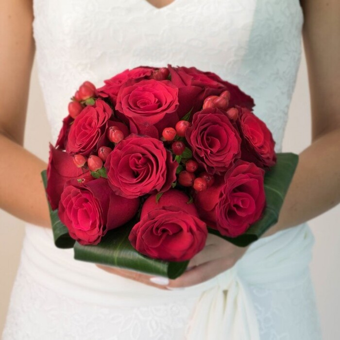 Presented in a waist-up composition, a Grove Park bride exudes quiet elegance, her white wedding dress adorned with softly detailed lace and flowing fabric that enhances the gentle light filling the space. The real star, however, is the superbly crafted bouquet resting in her hands-a compact sphere of flourishing, velvety red roses, their petals tightly layered and bursting with the colour of pure romance. Tucked amongst the roses, spiky clusters of luminous red hypericum berries add an energetic, jewel-like touch that sparkles, reminiscent of festive local traditions in Grove Park. The bouquet's foundation is fashioned from broad, glossy green leaves, meticulously folded to create architectural dimension and sharp contrast-a nod to the lush greenery seen throughout Grove Park itself. The bride's hands, perfectly manicured, cradle the bouquet with care and grace, dramatizing its central role in this moment of celebration. Soft, neutral backgrounds gently blur into the distance, sharpening focus on both the intricate craftsmanship and the emotional significance of the bouquet. This arrangement is a stunning example of timeless wedding florals, beautifully tailored for couples in Grove Park seeking to honour tradition while expressing deep love and new beginnings.