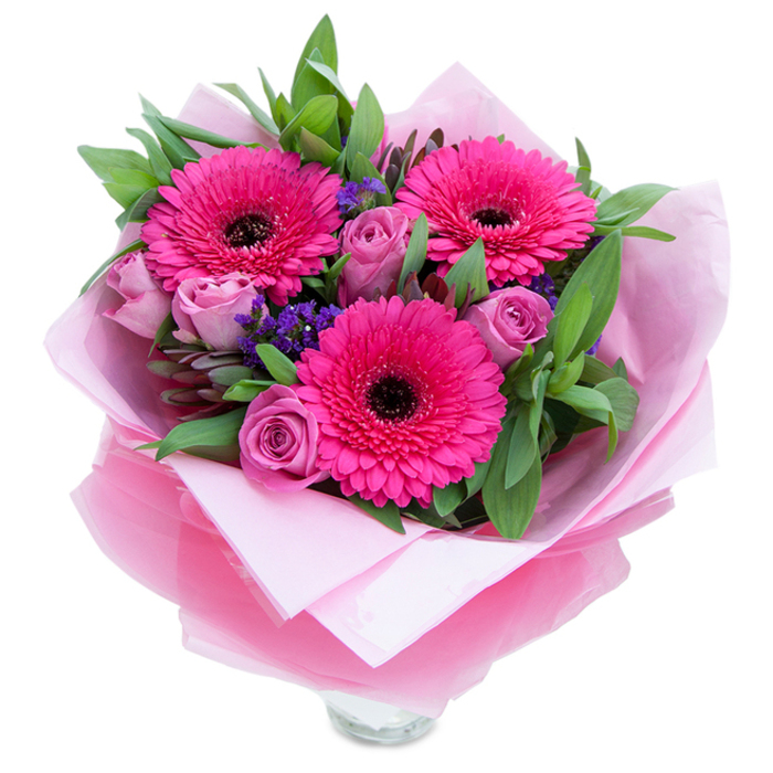 I picture this bouquet sitting on a small wooden table by a window overlooking Chinbrook Meadows, its tones a harmony of pinks and purples that read as both playful and refined. Dominating the arrangement are three vivid fuchsia Gerbera daisies - generous, round faces with countless petals that frame a deep, dark centre and draw the eye immediately. Soft lavender-pink roses nestle around them, their tightly furled petals suggesting freshness and a quiet romanticism. Between these principal blooms, sprigs of purple statice lend a papery, sky-lit texture that punctuates the pinks with cool depth. The greenery is broad and abundant, leaves like salal or eucalyptus giving the bouquet its structure and a subtle, green-note fragrance that balances the sweeter rose scent. The flowers are presented with care: an inner wrap of pale pink, matte tissue cushions the stems, and an outer layer of translucent, slightly crinkled fuchsia-pink cellophane adds a luminous sheen, folded with practiced hands so the bouquet reads polished and intimate. The base of a clear glass vase is visible beneath the wrap, indicating the piece is meant for immediate display and enjoyment. Bathed in gentle daylight the colours feel optimistic and warm - suitable for celebrating birthdays, anniversaries, or for brightening a friend's day. This is the kind of hand-tied arrangement that brings a touch of the garden indoors, made with the attention to form and scent you'd expect from a neighbourhood florist who knows Grove Park's quiet streets well.