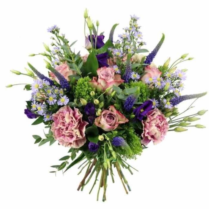 This hand-tied bouquet, captured against a clean white background, showcases a harmonious blend of colour and texture that feels very much at home in the calm, residential streets around Grove Park station. The flowers are arranged in a rounded, dome-like form, brimming with soft pinks, mauves, purples, and vivid greens. At the forefront are pastel pink roses, some fully open in generous, layered rosettes and others in earlier stages of bloom, their petals swirling tightly around deeper pink centres before fading gently to lighter, almost cream-toned edges. Interspersed with the roses are ample carnations in dusty rose and purple-mauve hues, their ruffled petals overlapping to create a dense, billowy texture that makes the bouquet appear rich and abundant. Rising through this softness, tall stems of deep lavender-blue Veronica introduce vertical lines and a gently tapering shape, which draws the eye upwards and adds a slight sense of movement. Among the more dominant blooms, clusters of purple lisianthus offer velvety, bell-shaped flowers, some more open and others in bud, deepening the cool, romantic palette. Threaded delicately between everything are fine sprays of aster-like blossoms in light lavender and periwinkle, each tiny, daisy-like head with a yellow centre giving a starry lightness to the whole design. The bouquet is framed by a generous layer of foliage, featuring bright, chartreuse-green textural elements that pop against the softer petals, and silvery-green eucalyptus leaves whose rounded shapes and muted tones soften the outline. At the bottom, neatly aligned green stems are clearly visible, securely tied in a natural-looking bundle that signals the bouquet is ready to be placed directly into a vase. The arrangement conveys a blend of modern elegance and romantic charm, suitable for gifting on birthdays, anniversaries, or simply to brighten a home in Grove Park with a sense of occasion.