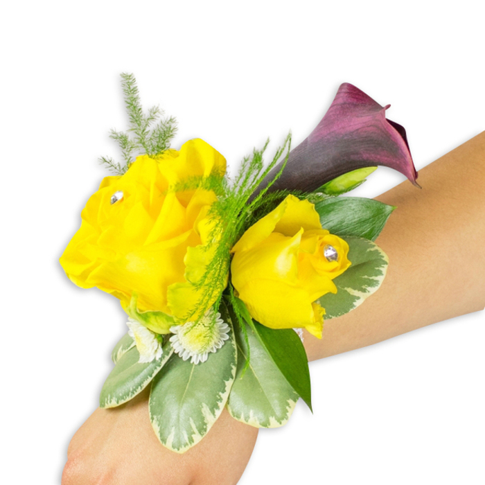 This finely crafted wrist corsage is displayed on a pale to light-medium forearm against a pure white backdrop, composed to show both the floral artistry and how it is worn. Two vivid yellow roses anchor the design: the left blossom is broader and more open, the right a slightly smaller, cupped bloom - each centre punctuated by a single sparkling clear rhinestone that lends a delicate, celebratory glint. Rising behind them is a solitary deep purple, almost black-cherry calla lily whose smooth, trumpet-shaped form provides a poised, sculptural contrast in silhouette and tone. Surrounding the trio are layers of texture: soft, airy asparagus fern threads that add lift; tiny white button-like filler blooms that give a dotted, cottage-like fullness; and sturdy variegated leaves with pale margins that introduce a cooling, waxy surface beneath the petals. A few solid dark-green leaves peek through to balance the composition. The corsage is fastened with a slim band so the blooms sit naturally on the wrist, arranged diagonally from the lower-left toward the centre-right of the frame to demonstrate wearability. The tactile impression combines velvety rose petals, the sleek calla surface, and feathery greenery, while the rhinestones catch the studio light with every slight movement. Thinking locally, this colour pairing - joyful yellow with dramatic plum - is a popular choice for weddings and formal nights out in Grove Park and nearby parks like Grove Park Nature Reserve or along Baring Road, offering a compact, spirited accessory that reads handcrafted and freshly prepared.