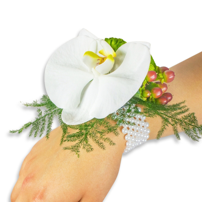 Presented on a pale wrist against a pristine white field, this wrist corsage combines classic materials and botanical finesse in a way we often craft here at Grove Park Florist. The star is a flawless white Phalaenopsis orchid, broad-petalled and satin-smooth, its central labellum painted with a vibrant yellow and a deeper golden-orange that deepens toward the throat, drawing the eye into the bloom's elegant architecture. Framing the orchid, fluffy sprays of asparagus fern create a soft, verdant bed - bright, clean green that provides lift and a feeling of lightness. Nestled to the upper right, a compact cluster of Hypericum berries introduces seasonal colour progression: pale green shifting to pink-rosy tones and finally to a warm cranberry red, each glossy orb catching light and echoing the orchid's warm centre. Small, ruffled lime-green accent leaves add a contemporary edge and tactile interest. The arrangement is secured with a multi-strand bracelet of tiny white pearls that wrap comfortably around the wrist; their pearly sheen brings a timeless, slightly vintage note that pairs beautifully with the modern botanical elements. The scent is gentle - a faint sweet hint from the orchid and fresh green notes from the fern - and the overall mood is romantic, composed, and wearable for weddings, proms or special anniversaries. Locally minded, this piece travels well across Grove Park and neighbouring Lee, Hither Green and Sundridge Park, available for local delivery and finished with the kind of careful assembly familiar to people who ask us for understated elegance close to home.