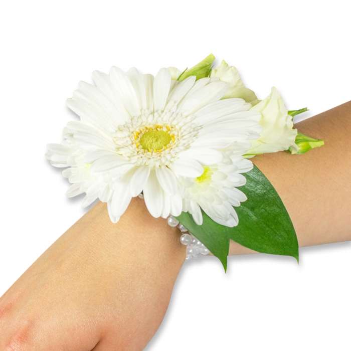 A classic white wrist corsage, artfully arranged by Grove Park Florist, is photographed on the forearm of someone with a light to medium skin tone against a neutral white backdrop. At its heart sits a large white Gerbera daisy, its soft, even petals radiating outward from a textured yellow-green centre that gives the piece a lively focal point. Surrounding this are clusters of small, snow-white chrysanthemums and tender cream lisianthus buds, layered to create a soft, cloud-like depth and a variety of delicate silhouettes. Rich, deep-green leaves-reminiscent of salal or ruscus-frame the cluster and extend slightly along the forearm, their glossy veins and smooth edges adding natural contrast to the pristine blooms. The flowers are secured on a subtle multi-strand pearl band that hugs the wrist with quiet elegance; the pearlescent beads catch the light and lend a touch of old-world charm. The composition evokes the cool, fresh scent of recently cut stems and a faint green tang that recalls early-morning markets near Grove Park station and walks through Chinbrook Meadows. This corsage is crafted to sit comfortably during long ceremonies or celebratory dances and is an ideal choice for bridesmaids, prom-goers, or graduates. Grove Park Florist prepares similar pieces for same-day delivery across Grove Park and neighbouring Hither Green and Downham, ensuring the blooms arrive fresh and ready to wear for the moment that matters.
