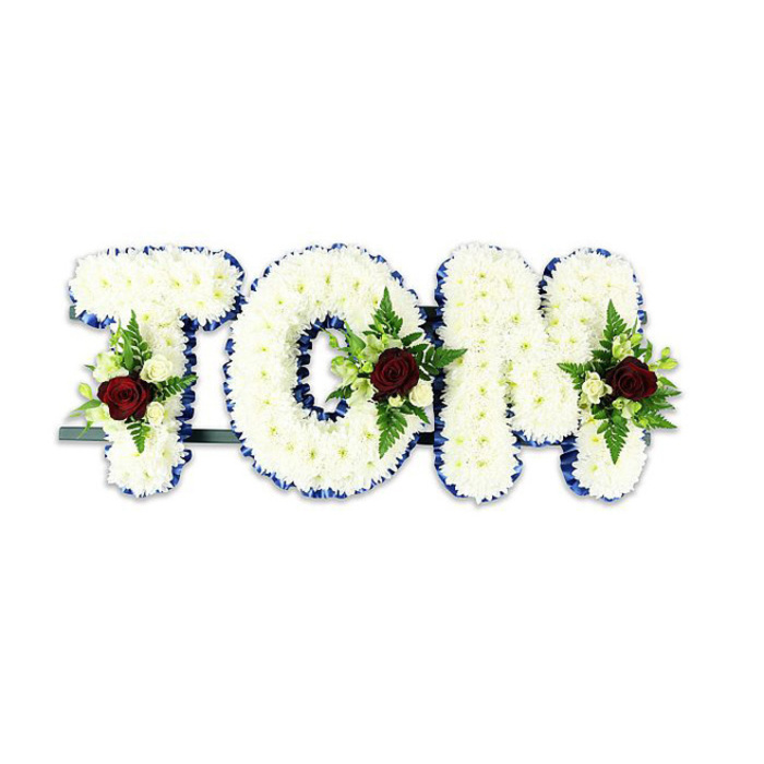 A striking, personalised memorial arrangement carefully spells the name TOM in large, sculpted letters formed from countless small white chrysanthemums, their petals layered to build a plush, tactile surface that reads as both pure and consoling. Each letter is sharply edged with a vivid royal blue ribbon that provides visual definition and a respectful, almost ceremonial border against the soft white masses. Within the central planes and lower sweeps of the T, O and M are small, symmetrical sprays: each anchored by a single, velvety deep red rose that stands as a symbol of enduring love and grief, accompanied by delicate ivory spray roses and fern fronds that introduce verdant highlights and graceful line. The craftsmanship is evident in the balance of colour and weight-the blue edging, white chrysanthemum bodies and concentrated red accents combine to form a composed and formal tribute. A slender, dark support bar is visible behind the base, the practical structure ensuring the letters remain upright when positioned on an easel in a chapel or funeral parlour. Photographed on a stark white background, the arrangement's textures, subtle shadows and neat lines are clear and readable. As a florist familiar with services across Grove Park and neighbouring Downham and Lee, I would recommend this style for memorials where a personalised name tribute is desired: it conveys dignity, comfort and a gentle, tactile sense of remembrance without excess ornamentation.