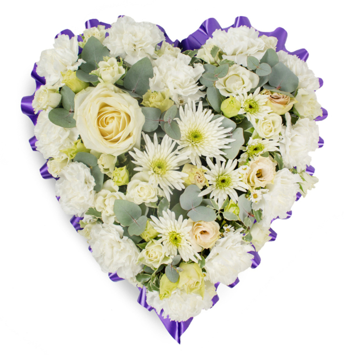 This image shows a large, heart-shaped sympathy arrangement resting against a pristine white background, its calm palette and careful structure inviting a moment of reflection. At the very centre sits a flawless creamy white rose, its petals delicately layered in a spiral that draws the gaze inward, like a quiet promise of enduring love. Around it spreads a blanket of white carnations, heavily clustered so that their ruffled edges touch and overlap, giving the heart a soft, cushioned appearance, almost like a pillow of petals. Among the carnations, bright white spider chrysanthemums reach out with fine, elongated petals, adding contrast and visual dynamism, while smaller daisy-style blooms with pale green or lemon centres introduce tiny points of colour and a subtle, cheerful brightness. Scattered here and there, ivory and cream rosebuds and lisianthus buds rest between the larger flowers, some partially open, suggesting the unfolding of shared memories over time. Interspersed throughout are slender strands and rounded sprigs of eucalyptus, their soft blue-green foliage weaving through the design to bring a hint of woodland freshness, reminiscent of walks through the green pockets around Grove Park and the Thames path. The entire heart is crisply edged with an opulent purple ribbon, carefully pleated and ruffled so it follows the curves with precision, framing the white blooms with a regal yet understated border. The overall composition is both structured and tender, as though expertly arranged by a seasoned Grove Park florist before being delivered to a church, crematorium, or family home. It communicates a deep sense of condolence, respect, and peaceful farewell, making it a fitting funeral tribute for families in Grove Park and the surrounding neighbourhoods.