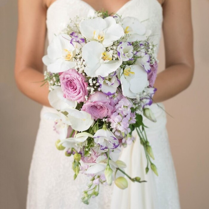 Gently illuminated by filtered natural light, a bride in Grove Park is captured from the mid-torso down, enveloped in the delicate textures of a strapless, lace-adorned wedding gown. Her arms tenderly cradle a cascading teardrop bridal bouquet curated with signature skill. Dominating this masterful creation, broad, pristine white phalaenopsis orchids display golden centers, radiating a quiet luxury amidst a medley of blush-toned garden roses and delicate lavender stock. Interspersed ruffled lisianthus, some accentuated by rich purple edges, add further complexity, their hues reflecting the spring colours seen around local gardens. Wisps of baby's breath and glimpses of green foliage offer a gentle breath of nature, softening the arrangement while preserving its immaculate flow. The bouquet's gradual descent mirrors the gentle curve of the bride's arms, creating a harmonious, organic silhouette. This bouquet, reflective of the refined artistry available at Grove Park Florist, brings together a romantic blend of tradition and modern design-perfect for weddings that wish to honour both the past and present spirit of the Grove Park community.