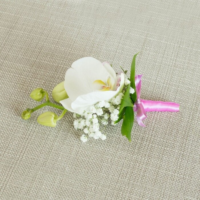 A refined orchid boutonniere, equally suited as a corsage, is presented lying horizontally on a softly textured, light beige linen-like background that feels calm and understated, much like a quiet corner of Grove Park in the early evening. Dominating the composition is a single, immaculate white Phalaenopsis orchid, its broad petals luminous and silky, opening with graceful curves that draw the eye straight to its centre. There, delicate markings of soft yellow and the faintest touch of pale pink add gentle warmth, giving the bloom a sense of depth and intricate beauty. Gathered snugly at the base of the orchid are clouds of tiny white baby's breath blossoms, their numerous, rounded petals forming a feathery, romantic halo that beautifully softens the strong silhouette of the main flower. From behind the blooms, a few slender, vibrant green leaves emerge, their clean lines and rich colour adding a fresh, natural contrast reminiscent of the greenery lining local paths and gardens. Extending to one side are several unopened lime-green orchid buds on a slim stem, promising future flowers and bringing a hint of movement and storytelling to the design. The stems are carefully bound together with a narrow band of bright fuchsia pink satin ribbon, tied in a small, precise bow whose sheen catches the gentle, even light. That vivid pop of pink against the white and green adds a celebratory accent, suggesting weddings, proms, or formal dinners held in and around Grove Park. The overall mood is one of polished simplicity and timeless elegance, as though prepared by a florist who understands how to let one exquisite orchid speak quietly, yet confidently, on a lapel or wrist.