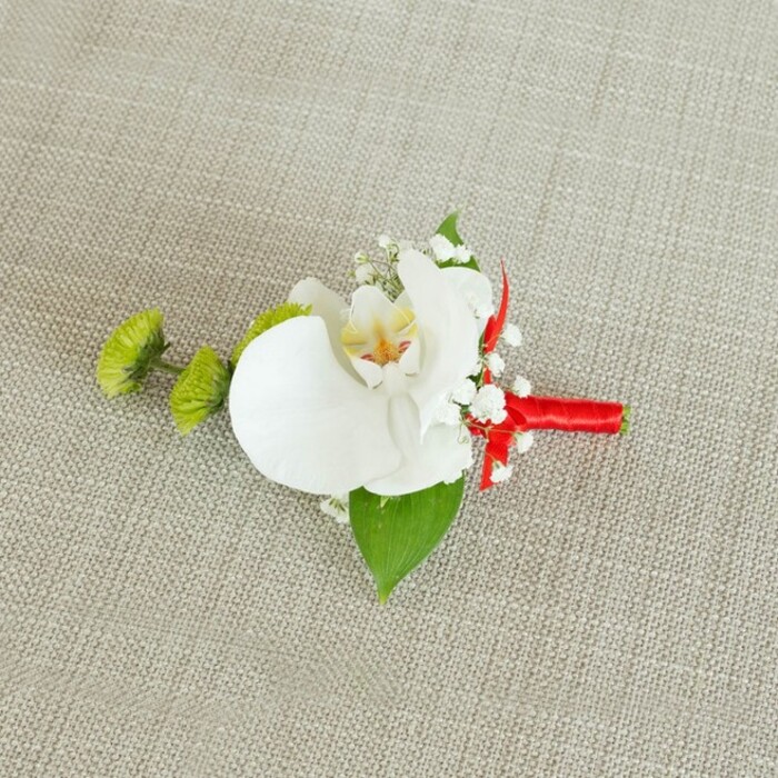 This photograph captures a meticulously crafted orchid boutonniere, composed with the quiet confidence of an experienced florist and set against a soft, light beige linen background whose slightly coarse weave adds a natural, tactile quality. At the centre, a pristine white phalaenopsis orchid bloom opens fully, each broad petal smooth and slightly curved, giving the impression of gentle movement even in stillness. The orchid's throat is detailed with subtle yellow tones that transition into hints of reddish-orange and light brown speckling, a small but striking focal point that rewards a closer look. Around this central flower, miniature white baby's breath blossoms nestle closely, creating an ethereal halo that lightens the composition and evokes the delicate haze of early spring mornings along the tree-lined streets of Grove Park. To the left of the orchid, two compact lime-green chrysanthemum pom-poms stand out, their tightly packed, fine petals forming a textured sphere that contrasts sharply with the orchid's smooth surface. A large, deep green leaf fans out beneath the main flowers, while a smaller, darker leaf peeks from around the ribbon, both adding depth and a sense of foliage freshly cut from a well-tended garden. The stems of the arrangement are neatly aligned and wrapped in a bold crimson satin ribbon, the fabric wound firmly and evenly to create a clean, cylindrical handle that ends in a modest, tidy knot. The red ribbon introduces a vivid stripe of colour that hints at romance and celebration, making this boutonniere perfectly suited to a groom or groomsman at a Grove Park wedding, perhaps on their way to a nearby church or local reception venue, where this understated yet luxurious piece would photograph beautifully on a dark suit lapel.