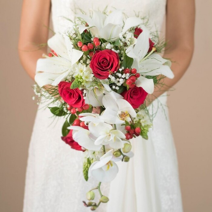 The image centres on a sumptuous, cascading bridal bouquet held in the gently clasped hands of a bride in a white lace wedding dress, her figure rendered in a soft blur so the flowers remain the star. The bouquet is crafted in a classic teardrop style, brimming with white and red blooms that feel both timeless and contemporary. At the heart of the arrangement, several large, trumpet-shaped white oriental lilies open wide, revealing bold stamens and delicately ruffled petal edges that catch the light. Their cool, creamy white tone contrasts beautifully with the rich crimson of velvety red roses, which are tucked throughout the bouquet in full, layered blooms that suggest devotion and deep affection. Flowing from the lower edge, slender sprays of white phalaenopsis orchids trail down in a loose cascade, their elegant, moth-like flowers and unopened buds giving the impression of movement, as if the bouquet were gently swaying while the bride walks. Interlaced among these focal flowers are tiny white blossoms, reminiscent of baby's breath, which add a romantic haze and soften the strong lines of the lilies and roses. Bright red berries, likely hypericum, peek between petals and foliage, bringing crisp pops of colour and a slightly rustic, seasonal charm that would feel perfectly at home at a Grove Park ceremony or a nearby riverside reception venue. Wisps of fine green fern-like foliage and deeper, sturdier leaves create a textured, layered base, lending support while keeping the overall arrangement light and airy. The bride's bare, lightly sun-kissed arms frame the bouquet, hinting at early autumn or late spring sunlight. Behind her, the background fades into a warm, neutral beige blur, much like the interior walls of a local Grove Park townhouse or small wedding venue, so that every curve of petal, gleam of berry, and thread of greenery can be appreciated as a work of floral artistry.