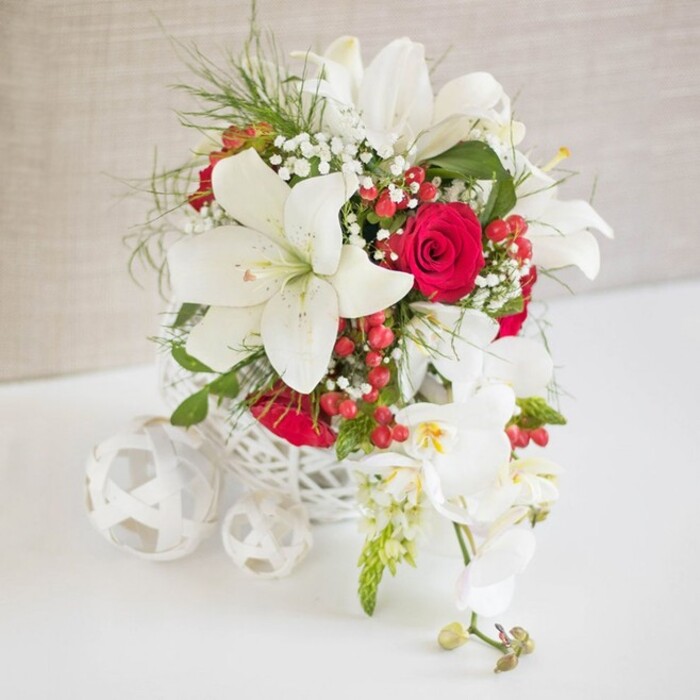 A luxurious white and red floral display rests on a crisp white surface, the entire scene set against a soft, light beige background that quietly recedes and allows every flower to stand out, as if styled for an intimate Grove Park celebration. The main bouquet is nestled in a round, openwork white basket, its intricate, lace-like weave suggesting hand-crafted artistry and adding an airy structure that supports the blooms without feeling heavy. Rising from this base, several impressive white Oriental lilies dominate the composition, their large petals opening in wide, star-shaped forms that reveal long stamens dusted with warm-toned pollen. Between these luminous lilies, velvety red roses are carefully placed, their deeply layered petals forming rich, cup-shaped blossoms that convey classic romance. Threads of fine white baby's breath run throughout, creating a light, clouded veil that softens the stronger shapes. Around and between the flowers, bright green foliage and fine, feathery greenery contribute energy and rhythm, adding a sense of living freshness reminiscent of nearby Grove Park and the leafy river paths. Small clusters of glossy red hypericum berries nestle among the stems, catching the light with tiny bursts of brightness that lend a subtle festive atmosphere, suitable for winter weddings or holiday dinners. To the right, a graceful strand of white phalaenopsis orchids cascades downward, each bloom perfectly formed with smooth white petals and a delicate yellow-and-soft-pink heart, followed by a few unopened buds that hint at future blooms. On the left side of the arrangement, three white woven spheres, echoing the basket's material, lie artfully on the table, tying the entire composition together with sculptural repetition. The bouquet exudes refined elegance and warmth, an ideal centerpiece for a wedding table, anniversary meal, or special event hosted in Grove Park's charming homes and local venues.
