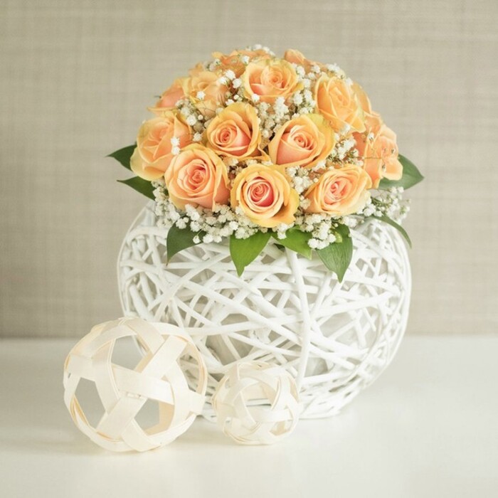 An elegant modern centerpiece is arranged with the precision and warmth you would expect from an experienced local florist, its soft peach tones standing out against a crisp, white setting. At the heart of the image is a domed bouquet of approximately two dozen roses in shades of apricot and light peach, each bloom nestled closely against its neighbour to create a full, almost cushion-like mound of petals. The roses show subtle variations in stage and shade: some are still tightly budded, with compact spirals encasing deeper tangerine hues, while others are just opening into layered rosettes, their outer petals fading to a creamy, almost shells-and-sand tone. Threaded generously among the roses are fine sprays of white baby's breath, the tiny clustered blossoms forming a gauzy veil that wraps around the arrangement like a soft cloud. Here and there, dark green leaves peek out beneath the blooms, lending contrast and suggesting the weight and freshness of the stems beneath the surface. The bouquet rises from a distinctive spherical vessel made of white woven strands, similar to string or thin wire, interlaced into an open, airy lattice that feels both artistic and minimal. Through the gaps in the weave, shadows and glimpses of the stems can be seen, adding depth and interest. This orb-like vase rests on an immaculate white tabletop, where two additional decorative spheres are placed in front-both crafted from the same white strand material. One is larger and more structured, faintly reminiscent of a stylised football, while the smaller one has a more irregular, sculptural form. Behind, a softly textured wall in pale beige or light greige creates a serene, uncluttered backdrop. The light is gentle and diffuse, with no harsh shadows, suggesting an interior setting in a calm Grove Park living room or a intimate local venue along Grove Park Terrace, perfect for a wedding, anniversary dinner, or stylish home decor display.