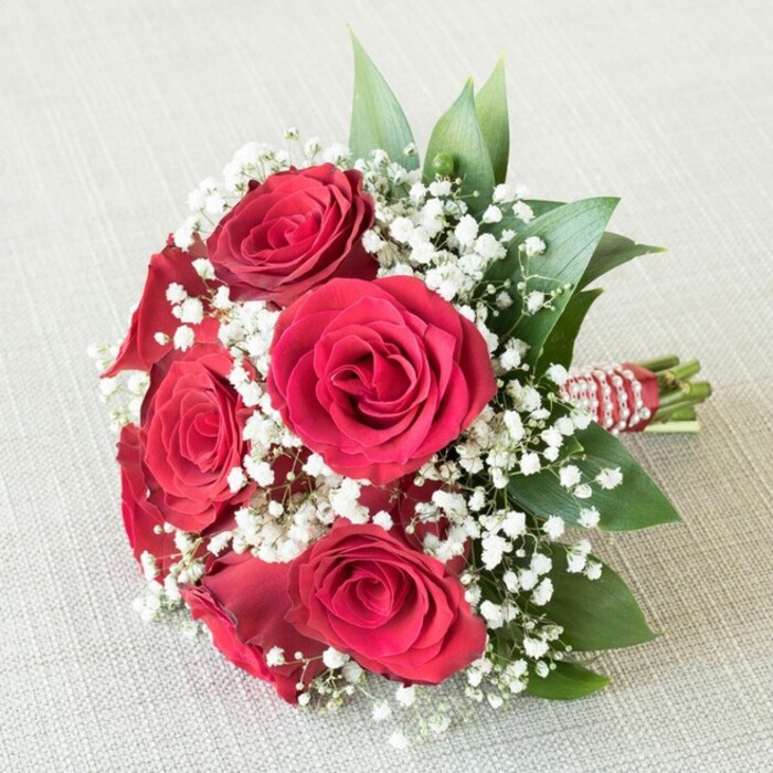 Representing the classic artistry of florists in Grove Park, this round, hand-tied bouquet features five to six lush red roses as its statement-each one blooming fully, the intricately layered petals unfolding with a rich, saturated crimson that embodies romance and grace. Among these, countless miniature white clusters of baby's breath (gypsophila) introduce a sense of lightness and airy movement, offering a dreamy counterpoint to the passionate intensity of the roses. The bouquet is defined by a frame of glossy, deep green foliage-perhaps ruscus or salal-which adds textural depth and freshness, reminiscent of the flourishing gardens and parks dotted throughout Grove Park. The sturdy stems, meticulously bound with a red and white striped ribbon and further adorned by a delicate spiral of shimmer-white pearls, showcase not just craftsmanship but the elegance often found at local ceremonies and celebratory gatherings. Presented against a softly lit, cream-toned material, the bouquet's lively colours are gently amplified, making every bloom and frill of foliage pop-perfect for illuminating a Grove Park wedding or anniversary celebration. Evocative of tender moments and the expert hands that assemble such arrangements, this bouquet serves as a heartfelt tribute to love and devotion within the local community.