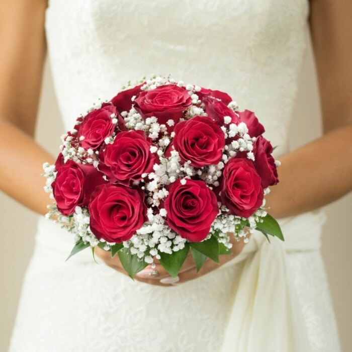Set against the softly blurred elegance of a neutral-toned Grove Park interior, a bride holds a domed bridal bouquet that signals classic romance and traditional artistry. Centre stage are a dozen opulent red roses, their petals undulating in velvet folds, creating an inviting depth and rich visual warmth evocative of celebratory days near Grove Park Common. Artfully woven throughout are delicate white baby's breath blossoms-gypsophila-contributing a lace-like, airy softness that tempers the bold crimson with ethereal calm. Peeking from beneath, fresh green foliage hints at the vibrancy of local nature, infusing a note of renewal and life. The bride's hands, manicured to perfection, cradle the bouquet confidently. Her wedding gown-perhaps adorned with lace or subtle embroidery-shimmers beneath the natural light, a gentle sash draping down the right for added movement. The background, softly faded to creamy beige, centers focus on the bouquet and the intricate texture of her attire. Every detail, from flower to fabric, reflects the refined skill of Grove Park Florist-a trusted presence for those planning a meaningful, elegant ceremony amid the area's tranquil streets and gardens. This image perfectly encapsulates timeless bridal beauty, becoming inspiration for wedding themes or local flower delivery alike.