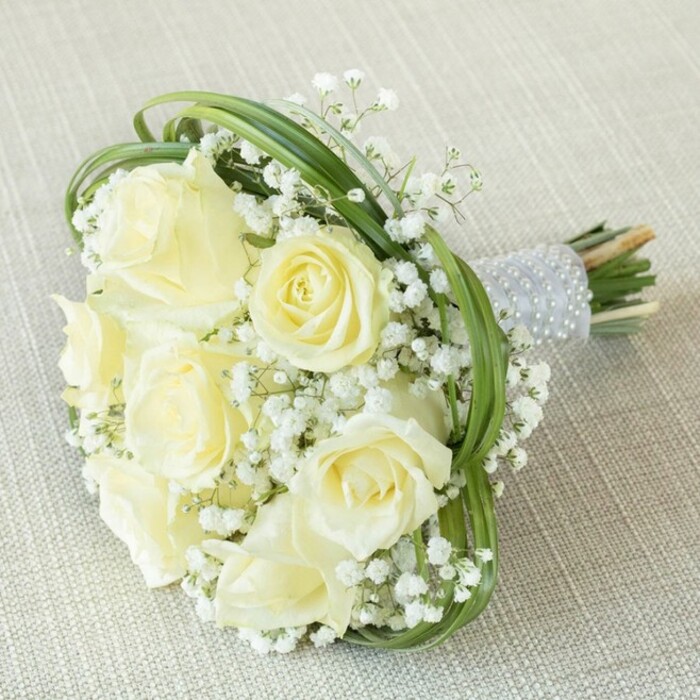 Against a gentle background of pale, textured fabric-a setting reminiscent of the soft, welcoming ambiences along Grove Park's residential avenues-this exquisite bridal bouquet makes a quietly powerful impression. Seven to eight luscious, fresh white roses, with petals unfurling in a creamy cascade, stand at the heart of the bundle, each conveying purity and gentle promise. Clusters of delicate, cloud-like baby's breath surround and support the roses, infusing the arrangement with a gossamer texture akin to lace glimpsed in a Grove Park window. Looped strands of vivid green bear grass form elegant arcs, delicately framing the flowers and echoing the leafy shapes seen near Chiswick House. The bouquet's stems, cut cleanly and gathered tightly, are embraced by a shimmering silver ribbon wrap, further adorned by neat rows of glossy pearl beads, lending a subtle shimmer in soft light. The organic simplicity of exposed green stems grounds the design in natural charm. Perfect for weddings, anniversaries, or memorable gatherings in Grove Park, this bouquet expresses timeless romance and the refined craftsmanship of a devoted local florist.