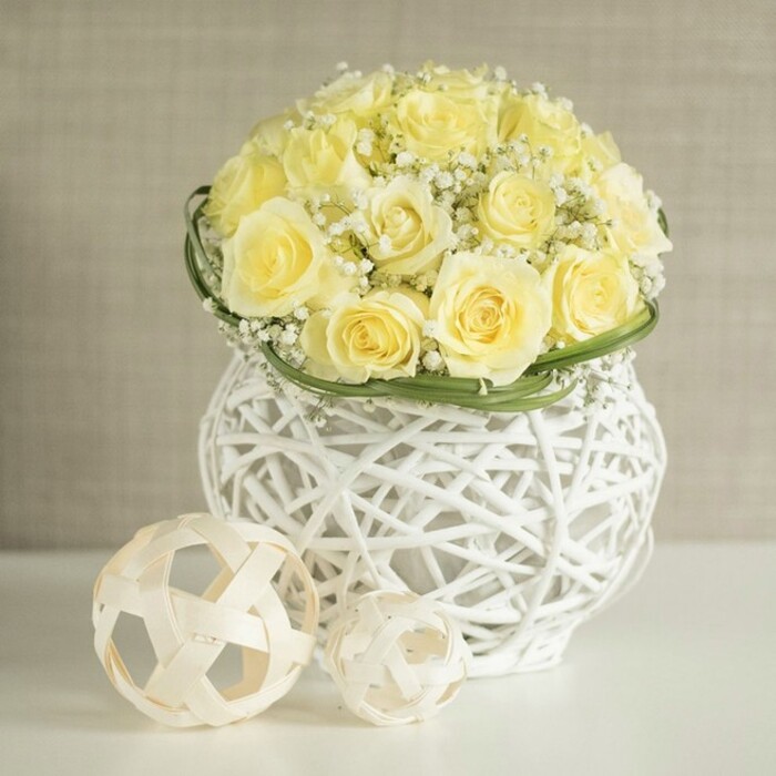 The photograph presents a graceful table arrangement anchored by a dome-shaped cluster of pale yellow roses, each bloom open at precisely the right moment, with petals unfurling in soft spirals of buttery colour. The roses are arranged so closely that they form a smooth, rounded canopy, with only the faintest glimpses of green stems hidden deep within. Interspersed throughout are airy clouds of white baby's breath, their myriad small blossoms resting lightly between the roses, introducing a delicate, cloud-like texture that softens and brightens the bouquet. At the very base of the flower dome, a narrow band of vivid green foliage or carefully woven leaves wraps tidily around the arrangement, adding structure and a modern, almost tailored finish that frames the blooms without distracting from them. The bouquet sits in a striking decorative vase, created from white, interlaced strands that imitate rattan or wicker, forming a gently curving silhouette that swells at the top and lower edge and draws in at the centre. The open structure of the vase allows plays of light and shadow to dance across the smooth white tabletop, where two light-coloured woven spheres are placed as complementary accents. These spheres, crafted from pale wooden or rattan strips, share the same open, geometric weave as the vase, reinforcing the theme of airy, natural textures. In the background, a soft beige or light grey wall fades into blur, reminiscent of a quiet corner in a Grove Park townhouse, ensuring that the flowers and their sculptural container remain the undeniable focus. The overall impression is one of understated luxury and gentle joy, perfectly suited for wedding breakfast tables at local Grove Park venues, stylish home gatherings near Chislehurst and Lee, or as a serene centerpiece delivered by Grove Park Florist to families celebrating milestones together.