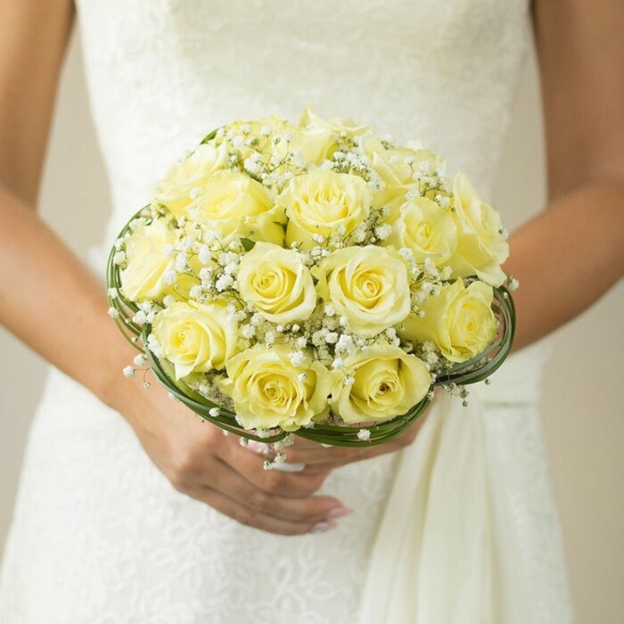 A bride's bouquet from Grove Park is revealed in exquisite, close-up detail, encapsulating the intimate artistry of West London's revered florists. The soft, harmonious palette begins with a mass of lush yellow roses, each flower perfectly in bloom, petals delicately unfurling to create a look of plush abundance. Nestled amongst these roses are tiny, star-like clusters of pure white baby's breath, their gentle form imparting lightness and evoking the dreamy simplicity of a summer's morning along Grove Park's riversides. At the bouquet's foundation, thin bands of rich green foliage or decorative wire are entwined, their subtle curves offering elegant structure and paying homage to the neighbourhood's well-kept gardens. The bride's steady hands are visible, nails pale and refined, as she supports her bouquet with an air of serene anticipation-a simple wedding band shining subtly in the light. The textural contrast continues into the background as the white wedding gown-perhaps adorned in lace or fine embroidery-fades softly, allowing the dazzling flowers to take centre stage. This scene is the epitome of celebration and emotion, the bouquet embodying romance and craftsmanship for Grove Park couples looking to embrace tradition with a fresh, sophisticated twist.