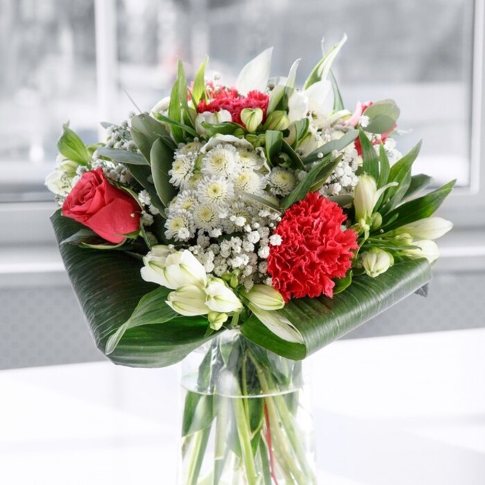The image shows an elegant, contemporary bouquet displayed in a cylindrical clear glass vase, positioned on a fresh white surface that reflects the natural light. Inside the vase, the green stems are neatly trimmed and gathered in a tidy spiral, visible through the water and revealing the skilled hand of a professional florist. Rising from this base is a lush, full arrangement built on the timeless contrast of red and white. Deep red roses, their petals soft and velvety, open in graceful swirls that catch the light, while a prominent crimson carnation near the front brings intricate texture, each finely ruffled petal adding depth and movement. Scattered among these passionate reds are refined white blooms: alstroemeria with slim, tapering petals delicately flecked with speckles, and several smooth, ivory lily buds still closed, poised to unfurl in the days ahead. Wisps of gypsophila, or baby's breath, thread throughout the bouquet, their tiny white flowers forming a light, airy veil that softens the stronger shapes and creates an almost misty halo. Around the base, broad, glossy dark green tropical leaves are wrapped and curled into a structured collar, lending the design a polished, architectural feel. Additional foliage in eucalyptus-like tones and pointed, lance-shaped leaves extends upward and outward, creating layers of green that frame the flowers and add a sense of natural abundance. The softly blurred, bright backdrop hints at daylight streaming through a window, perhaps overlooking one of Grove Park's leafy gardens. The arrangement feels both modern and romantic, perfectly suited as a centrepiece for a family gathering, an intimate anniversary dinner, or a thoughtful delivery to share gratitude and affection with someone special nearby.