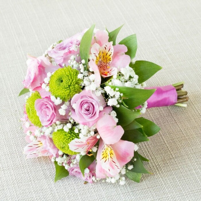 A striking, hand-tied bouquet crafted by Grove Park's seasoned florists is elegantly laid on a subtly textured light beige fabric, evoking the sophisticated tables and historic venues of the area. The arrangement is a medley of pastel pink and lavender roses-petals gently spiraled for classic romance-that sit in harmony with vibrant lime-green button chrysanthemums, whose dense, rounded forms add both visual and tactile depth. Interspersed are refined pink alstroemeria, petals washed in blush with yellow and burgundy speckles, opening wide to reveal a charming, painterly effect reminiscent of botanical artistry seen at Grove Park events. Wispy sprays of white gypsophila float between the blossoms, imbuing the bouquet with lightness and a hint of early morning mist often found on Chiswick Lane. Rich dark greenery frames and supports the floral design, grounding the colors and echoing the lush parks nearby. The handle is neatly finished with a vibrant magenta satin ribbon, tied just tightly enough to hold the bouquet but leaving the stems exposed, offering an authentic, organic finish and a sophisticated nod to contemporary bridal trends. With its radiant colors and careful composition, this bouquet is perfectly suited for Grove Park bridesmaids, anniversary gifts, or as a stunning table centrepiece-each element reflecting the neighbourhood's dedication to beauty and celebration.