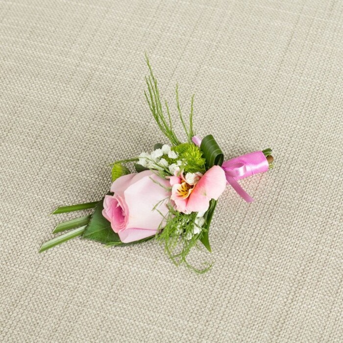 A carefully composed boutonniere, positioned against a light, natural linen, reflects both Grove Park's peaceful nature and the skilled hands of a local floral designer. The focal point is a youthful pink rosebud, petals tightly coiled in a gentle spiral and gradating from a vibrant rose-core to whisper-soft edges. Alongside, peach-pink alstroemeria reveals its luminous petals with subtle yellow glimmers and patterned throats, reminiscent of the delicate blooms found in the gardens off Hartington Road. Baby's breath acts as a floating accent, with minute white blossoms scattered through the boutonniere, while miniature ball-shaped green blossoms add a zest of life. Spiky, fern-like sprigs and broader, deep-green leaves compose the rest, adding verdant volume and a structured backdrop. A wide emerald leaf folds around the base, securing the layout and lending a modern edge. The stems are bound with a striking fuchsia satin ribbon, expertly tied into a sharp bow that catches the light and draws the gaze. Resting on the oatmeal-toned, slightly coarse linen, the inspired arrangement stands out with classic yet contemporary vibrancy. Worn at a Grove Park wedding, prom, or celebratory gathering, it promises an air of refined festivity and artisanal charm-perfect for commemorating meaningful occasions in the local tradition.