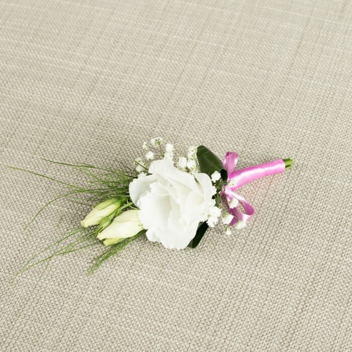 A small, meticulously crafted boutonnière is laid diagonally across a pale beige, textured fabric backdrop, the weave clearly visible like a fine linen runner you might see on a Grove Park dining table prepared for a celebration. The central focus is a fully opened white lisianthus bloom, its petals soft and gently frilled, spiralling outward in generous layers that form a rounded, cup-like flower head. The innermost petals glow faintly cream against the pure white outer edges, hinting at depth and freshness. Nestled just below and to the left of this main flower are two elongated lisianthus buds, still closed, their slim forms blending creamy white and pale green, suggesting the promise of flowers yet to open as the day unfolds. Interwoven among the main bloom and buds are delicate sprays of baby's breath, with tiny, cloud-like white flowers that create a fine, lacy mist around the focal lisianthus, adding a sense of softness and romance often requested for local Grove Park weddings. From behind the flowers, slender, grass-like leaves in a deep, cool green stretch out towards the lower left corner, creating gentle lines of movement and drawing the eye outward. A wider, darker green leaf rests just behind the central bloom, providing a subtle, supportive backdrop. At the base on the lower right, the stems are trimmed neatly and wrapped with a bright fuchsia satin ribbon, tied into a tidy little bow whose smooth, glossy surface catches the light and introduces a bold splash of modern colour. The lighting is even and calm, revealing each texture-from the woven fabric and satin sheen to the velvety petals-as if the piece is waiting on a dressing chair before being pinned on for a ceremony or prom night near the quiet streets and open green spaces of Grove Park.