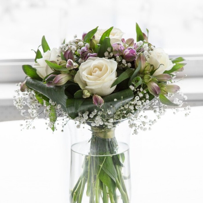 In this image, a softly luminous bouquet of creamy white roses, lavender-hued alstroemeria, and delicate baby's breath is presented in a clear, cylindrical glass vase, as if placed on a windowsill in a quiet Grove Park kitchen or sitting room. The light appears natural and gently diffused, washing over the arrangement and highlighting the plush texture of the fully opened roses, whose petals unfold in generous, symmetrical layers that suggest classic romance and sincerity. Around these focal blooms, clusters of alstroemeria in muted purples and soft lilacs provide movement and variety, their elongated buds and just-opening flowers adding a sense of unfolding possibility, as though the bouquet is slowly coming to life over the course of a day. Tiny, cloudlike tufts of gypsophila nestle between the larger flowers, catching the light and lending a shimmering, lace-like quality that helps the arrangement feel both full and airy at once. Deep, glossy green leaves form a supporting frame, curving outward and upward, while finer foliage threads through the interior, giving the design a rounded, balanced silhouette that appears meticulously constructed yet naturally relaxed. Through the vase, the stems are clearly visible, neatly trimmed and arranged in a loose spiral, their fresh green tones intensified by the clean water that fills the vessel. The combination of whites, greens, and soft violets creates a tranquil, hopeful mood, reminiscent of early spring mornings walking towards the river from Grove Park. This bouquet would feel at home as a wedding anniversary gift, a graceful token of sympathy, or a simple gesture of appreciation delivered to a friend's doorstep, its gentle colour story and thoughtful composition reflecting the steady expertise of a trusted neighbourhood florist.