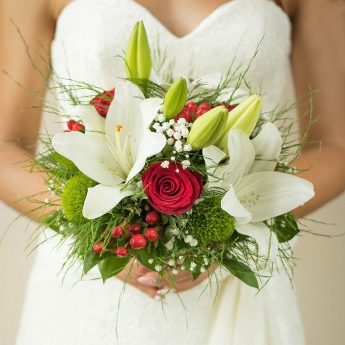 Bathed in the gentle light of a Grove Park celebration, a bride stands gracefully adorned in an elaborately textured white gown, clutching a wedding bouquet that masterfully blends tradition and contemporary flair. Pristine oriental lilies, lush and bright, dominate the display, each blossom opening with a golden flash of stamens that shine against the soft, flowing fabric. Fresh, unopened green lily buds are nestled amidst the blooms, adding an element of anticipation-a nod to all the tomorrows that await. At the heart, a singular deep red rose swirls with passion, its tightly layered petals drawing the eye and evoking the classic romance found at local Grove Park venues. Surrounding it, delicate sprays of baby's breath lend a dreamy, airy touch, while vibrant red hypericum berries add cheerful pops of colour that echo the warmth of the local community. Playful green button dianthus and a cascade of feathery and broad-leaf greens contribute texture and fullness, recalling the verdant landscapes along Strand-on-the-Green. The bride's softly clasped hands and the backdrop of her detailed gown invite intimacy and reverence for this pivotal moment. Carefully assembled by Grove Park Florist, this bouquet celebrates the unity of love and locality, making it an ideal choice for couples who want a wedding style that's both timeless and unmistakably rooted in West London's charm.
