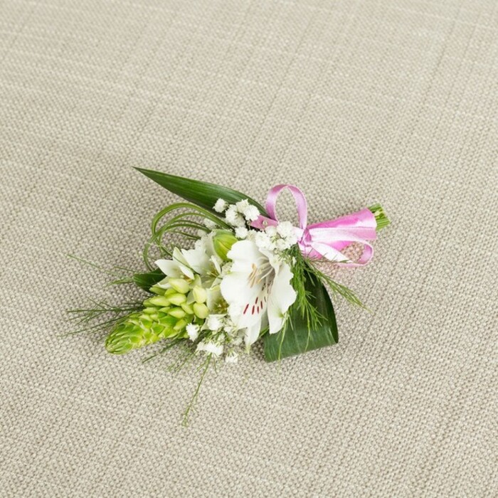 An artfully composed boutonniere or small corsage is displayed on a softly textured background of light beige fabric that resembles finely woven linen, the neutral tones echoing the calm residential streets of Grove Park. At the centre of the arrangement sits a single, open alstroemeria bloom in pure white, its petals gently flaring outward with a faint, velvety sheen. Near the throat of the flower, reddish-brown speckles form natural brushstrokes, lending depth and character to the otherwise pristine petals, while the stamens stand delicately within, suggesting the flower's natural fragrance and vitality. To the left of this focal bloom rises a compact, conical cluster of lime green buds, tightly layered and reminiscent of an Ornithogalum or ginger-like spike; its fresh, almost juicy colour adds a modern twist to the traditional boutonniere style. Threaded through the design, fine stems of baby's breath hold clusters of tiny, cloud-like white blossoms, softening transitions between the main flowers and adding a romantic, misty lightness. Supporting everything is a base of rich greenery: broad, dark green leaves with pointed tips create a strong, stable foundation, while slender blades of bright green grass arch and curl around the composition, suggesting movement and drawing the eye along gentle, looping lines. At the right edge, the stems are neatly bound with a bright, satin-finish pink ribbon, fashioned into a small bow whose glossy surface catches the light and adds a playful, celebratory accent. The lighting is even and flattering, allowing textures-from the crisp petals to the delicate baby's breath-to be clearly seen without harsh contrast. As a whole, this fresh floral accessory feels perfectly suited to weddings, proms, and formal occasions around Grove Park, prepared with the kind of care and attention to detail you'd expect from a long-established local florist.