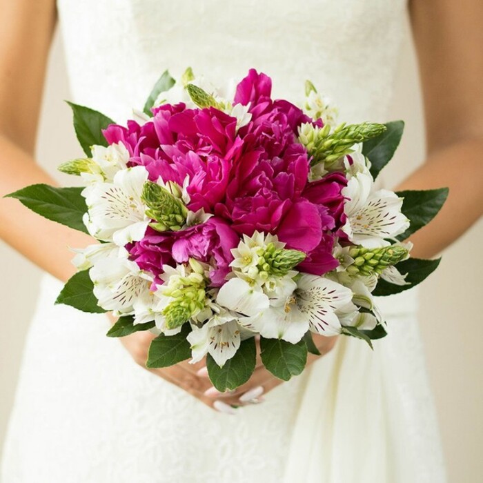 The photograph focuses closely on a rounded bridal bouquet held just at the bride's waist, set against the soft, slightly textured backdrop of a white wedding dress. At the centre, sumptuous peonies in rich fuchsia and magenta tones create a dense, velvety core, each bloom layered with generous, ruffled petals that tumble over one another in a luxurious swirl. Threaded thoughtfully among these dramatic flowers are white alstroemeria, or Peruvian lilies, their more slender petals opening gracefully and revealing delicate maroon speckles and fine lines in their throats, as if lightly dusted with ink. Sprinkled throughout, vivid green buds and spiky textural blooms push upward and outward, introducing a sharp contrast in both shape and colour that keeps the bouquet from feeling too traditional. Around the perimeter, wide, dark green leaves with a subtle sheen cradle the arrangement, offering a calm, natural frame that evokes a garden edge along a Grove Park terrace or a leafy pathway leading down to the Thames. The bride's hands appear at the base, fingers gently wrapped around the stems, nails finished with a pale pink polish that quietly echoes the bouquet's romantic palette. The background is softly blurred, likely more of the gown's fabric, so the eye rests on the interplay of bright fuchsia, clean white, and varied greens. There is a sense of joyful celebration yet also intimacy, as if the bouquet has been captured in a quiet pause before stepping out onto a sunlit Grove Park street, ready for photographs near the local churchyard or a favourite corner of the park. Every petal, marking, and leaf seems chosen to convey warmth, personality, and the freshness of a wedding morning in West London.