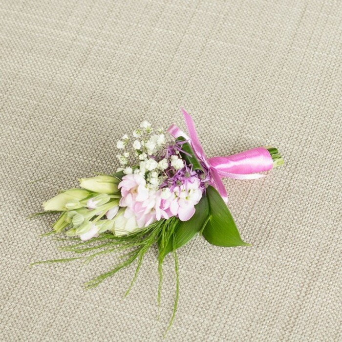 A single, meticulously composed boutonniere or small corsage rests on a softly textured, light greige linen backdrop, the fabric's fine weave and neutral tone providing a calm, understated stage that allows the flowers to shine. At the core of the arrangement, several full-petalled, light pink blooms sit closely together, their ruffled petals layered like miniature garden stocks or delicate cottage flowers you might see spilling over a fence near Grove Park station. Their colour transitions in subtle gradients from almost white at the outer edges to a more saturated blush in the centre, giving depth and softness all at once. Interlaced among them are clusters of tiny white baby's breath blossoms, each forming a small, cloud-like puff that adds texture and a sense of airiness. Touches of lavender and light purple florets peek from between the pink petals, adding a cool counterpoint that enriches the pastel palette without overwhelming it. On the left-hand side of the design, a graceful line of elongated, pale green to creamy white buds, likely freesias or similar slender blooms, extends outward, suggesting fresh, unopened flowers ready to reveal their fragrance later in the day. The floral cluster is anchored by a base of lush, broad green leaves, their polished surfaces offering a deep emerald contrast, while wisps of fine, grass-like greenery splay gently from the edges, hinting at something naturally gathered from a local Grove Park garden. The lower stems are neatly bound in a striking fuchsia satin ribbon, tied into a precise bow whose glossy loops and short tails introduce a playful yet polished accent. Soft, even lighting captures the freshness of each element, evoking the sense of a carefully prepared wedding or prom accessory, perhaps being readied by Grove Park Florist for same-day delivery to a nearby celebration.