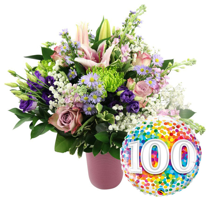 On a pure white background, a lavish, 100-themed floral arrangement captures the essence of a once-in-a-lifetime celebration, as though prepared for a beloved Grove Park resident reaching their centenary. The flowers are gathered in a soft, dusty-pink vase with a subtle vertical ribbed pattern that adds understated texture and sophistication. The bouquet itself is full and rounded, overflowing with blossom and foliage. Standing tall at the centre are light pink lilies, some fully open into broad, star-shaped flowers with delicately arching petals and visible stamens, others still in bud, promising more blooms in the days ahead. Encircling them are roses in shades of gentle blush and deeper mauve, each rose head unfolding in luxurious, velvety layers that suggest both romance and dignity. Deep purple lisianthus add a rich, jewel-like contrast, their slightly frilled petals curling inward around the centres. Dotted between the larger flowers, dainty lavender asters with yellow eyes lend a fresh, meadowy feeling, as if gathered from a quiet green space along the river near Grove Park. Small lime green button chrysanthemums provide lively accents, their tightly packed petals forming neat, spherical flower heads that complement the looser shapes around them. Throughout the bouquet, fine clouds of white baby's breath float delicately, softening edges and filling out the composition without overwhelming it. Various shades of dark green foliage-some leaves long and slender, others broader and more robust-frame the mix of pinks and purples, grounding the design. To the lower right, just in front of the vase, a round foil balloon with a glossy finish completes the celebratory scene. Its surface is blanketed in colourful confetti dots in red, orange, yellow, green, blue, and purple, creating a sense of fun and festivity. In bold contrast, the large white numerals 100 appear in the centre with a subtle shadow, making the milestone unmistakable and transforming the entire image into a joyful tribute to a hundred years of life and love in Grove Park.
