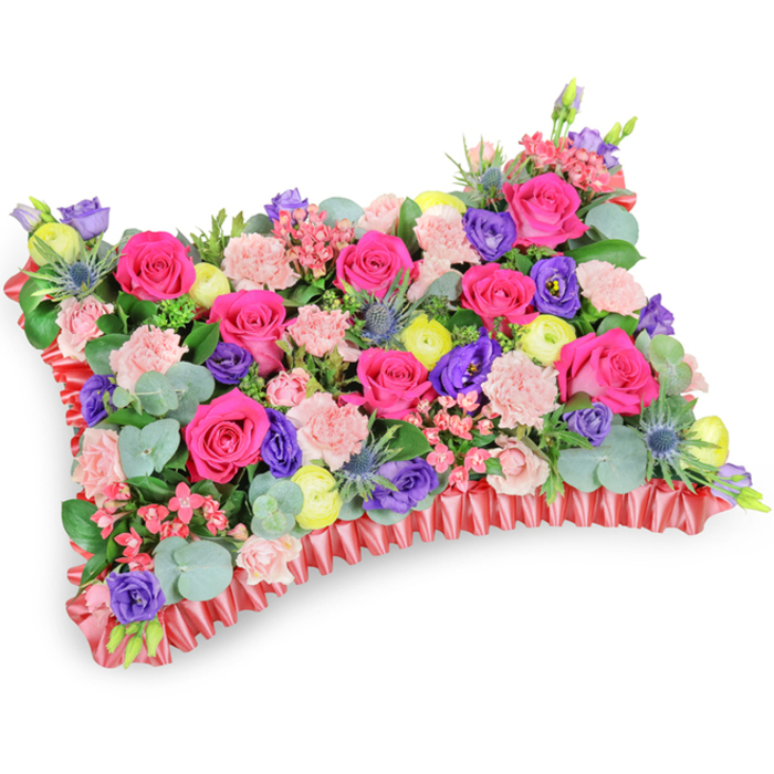 From a modestly elevated oblique perspective against a white backdrop, this densely composed pillow-style floral tribute presents as a lush, jewel-bright tapestry of blooms. Dominant hot pink roses sit like small cushions themselves, their satiny petals folding inward and catching soft highlights; intermingled are pale pink carnations that add a feathered, ruffled texture across the surface. Deep purple lisianthus introduce cool, velvety pockets of contrast while buttery lemon-yellow ranunculus appear as joyful, layered accents that brighten the composition. Blue-tinged eryngium thistles punctuate the arrangement with spiky, architectural interest, offering a steel-blue coolness against the predominantly warm palette. Sprays of fine pink fillers and the rounded silvery leaves of eucalyptus drift through the massed flowers, their matte, aromatic presence giving the pillow a restful, garden-fresh character. The whole sits on a decorative border of rich red, pleated satin ribbon that frames the rectangle with a soft, ruffled edge-an elegant finishing touch that clarifies the pillow silhouette and elevates its ceremonial purpose. The floral mechanics are dense and supportive beneath, the arrangement crafted to sit neatly on a casket, pedestal, or graveside display. The colour story-fuchsia, violet, lemon yellow and blush-leans toward celebration and cherished memories, softly easing grief with warmth. This is the kind of thoughtful memorial cushion often requested by families across Grove Park, Lewisham and neighbouring Lee, chosen to honour a life with beauty, scent and tactile comfort.