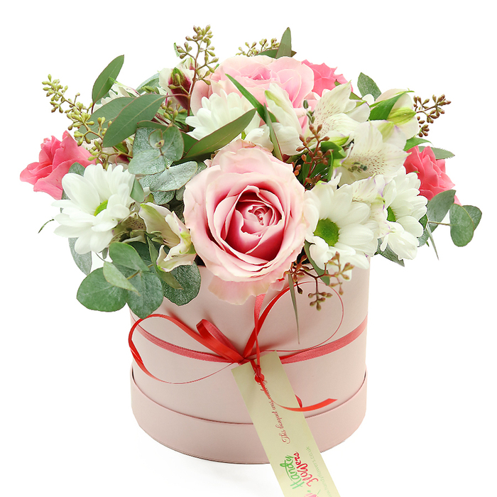 This image shows a beautifully composed floral hatbox arrangement, placed squarely against a bright white background that highlights its elegant colour palette. The box itself is round and cylindrical, finished in a soft, pastel pink that appears smooth and matte to the touch. Around its lower half runs a slim coral-red satin ribbon, meticulously tied into a small, refined bow. A rectangular cream tag with subtle green handwritten-style lettering hangs from the knot, creating the impression of a bespoke gift prepared by an experienced Grove Park florist, perhaps destined for a townhouse off Sutton Court Road or a cosy flat near the station. Emerging from the open top of the box is a generous dome of flowers that gently overflow the edges, giving an impression of fullness and natural ease. Several large roses, in shades of pale pink with hints of deeper fuchsia at their centres, form the heart of the arrangement; their spiralling petals layer upon one another to create a soft, romantic focal point. Scattered throughout, white daisy-like chrysanthemums with bright green, almost chartreuse centres provide contrast and a lively, cheerful energy. Elegant white alstroemeria blooms add length and lightness, their inner petals feathered with fine speckles that reward a closer look. Nestled among these are smaller, more intense deep pink blossoms - spray roses or lisianthus - which lend rich colour accents and visual depth. A mix of foliage, including rounded silvery eucalyptus leaves, darker green leaves, and a few delicate stems tipped with tiny buds or seed pods, frames the flowers and softens the edges. Overall, the arrangement conveys refined sentiment, making it perfect for anniversaries, Mother's Day, or thoughtful thank-you gifts around Grove Park, blending modern hatbox styling with classic floral beauty.