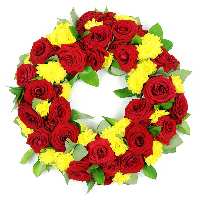 A large, circular floral wreath designed for funerals or memorial services is presented on a pure white background, making the colours of the flowers appear especially vivid and clear. The wreath is completely covered with blooms, forming a deep ring of petals without any obvious gaps. Deep red roses dominate the design, their velvety petals unfolding in tight, elegant spirals that create soft shadows and a sense of depth. Shades of red shift subtly from a rich crimson at the heart of each flower to a more intense, almost burgundy hue towards the outer edges of the petals. Between these roses, abundant clusters of bright yellow chrysanthemums have been placed to create soft cushions of colour. The chrysanthemums are made up of many fine, narrow petals arranged around a small central disc, giving each cluster a delicate, feathery texture that contrasts with the smooth rose petals. Surrounding and interwoven among the flowers is a base of fresh greenery. Broad, dark green leaves with a glossy finish catch the light, while some lighter, slightly silvery leaves-suggestive of eucalyptus-add a gentle cool tone and a more natural, garden-like feel. The wreath forms a perfect, unbroken circle, with a clean open centre that allows the viewer to see clearly through the middle of the arrangement, symbolising eternity, unity, and continuous remembrance. The lighting is bright and even, enhancing every detail of the craftsmanship, as if the wreath has just been finished by experienced hands at Grove Park Florist, ready to be delivered for a thoughtful farewell at a local chapel, cemetery, or riverside memorial gathering in Grove Park.
