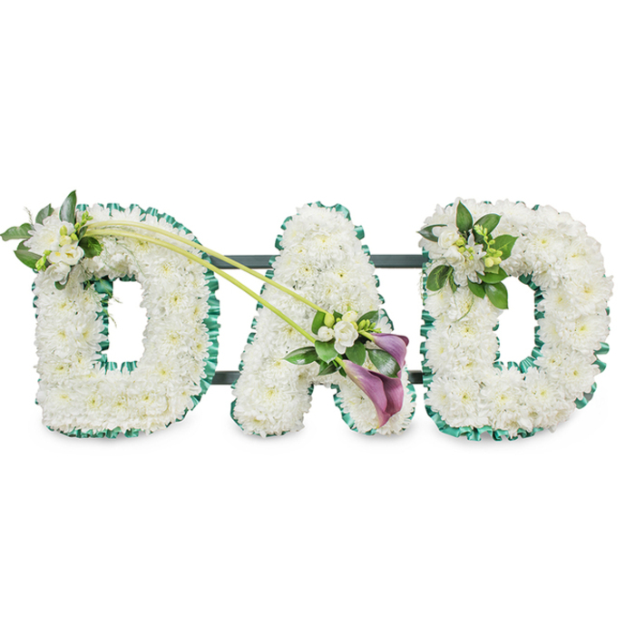 In this serene funeral arrangement, the word DAD is spelled out in large, upright floral letters, standing firmly on a hidden dark base that ensures they remain aligned as a single tribute. Each letter is built on a sturdy frame and then entirely covered with dense clusters of white chrysanthemums, the flowers so tightly packed that they form a smooth, cushion-like surface with a pleasing, uniform texture. Their clean, white petals symbolise remembrance and peace, making the piece particularly suited for a father's farewell. Along the outer edges of each letter, a rich, deep green satin ribbon is artfully ruffled, creating a delicate fringe that highlights the contours and introduces a fresh, natural contrast, reminiscent of the green avenues and quiet gardens around Grove Park. The first D is adorned at the top left with a small, elegant cluster of white freesias interwoven with glossy foliage, the delicate blooms suggesting a whispered message of love. Two slender green stems extend from this accent, sweeping diagonally toward the central A like a gentle arc. At their meeting point, close to the lower right side of the A, sits a graceful focal spray composed of two deep purple calla lilies, whose sleek, trumpet-shaped blooms rise from a base of white freesias and lush greenery. This rich colour accent adds depth and a slightly contemporary touch to the traditional letters. The final D features a more modest echo of this detailing on its upper right curve, with a neat cluster of freesias and foliage that completes the symmetry. The tribute feels quietly personal, an arrangement a Grove Park family might place by the coffin or at the crematorium to honour the memory, strength, and kindness of their dad.