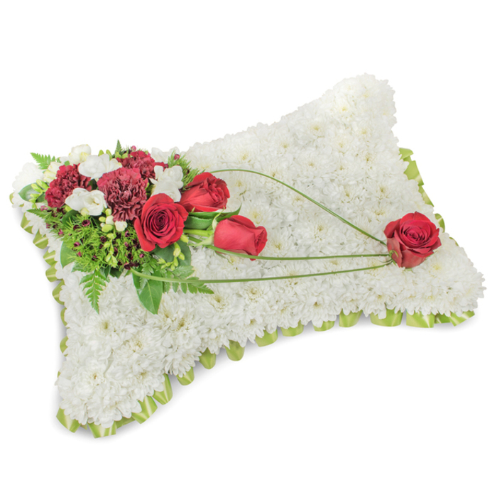 A poignant rectangular floral pillow, shown on a clean white surface so the arrangement's structure and texture are clearly visible: the whole form is meticulously covered with hundreds of tiny, pure white chrysanthemum blooms, their dense heads building a soft, cushion-like plane that evokes rest and serenity. Along the edge a subtle lime-green satin ribbon or fine foliage border is visible, lending a neat, finished silhouette. A rich corner spray at the upper left provides the emotional focus-several dark crimson roses, some in tight bud and others unfolding, set alongside deep burgundy carnations whose ruffled petals add sculptural contrast. Tiny clusters of white freesias or small spray chrysanthemums are woven through the reds, bringing brightness and a delicate scent. A tapestry of green supports the flowers: feathery fern fronds, glossy leaves and slender accent stems that create shadow, texture and a sense of life. From that corner the design carries a graceful movement-three arcing lines of bright green bear-grass or decorative ribbon sweep across the white chrysanthemum field toward the lower right, where a single, perfectly formed red rose rests like a final, reverent gesture. The lighting is soft and even, revealing gentle petal edges and the cushion's subtle contours without dramatic highlights; the overall mood is one of calm dignity and compassionate care. This classic memorial cushion, often chosen for funeral services, wakes or remembrance displays near Grove Park's community halls or nature reserves, speaks of respect, lasting affection and careful florist craftsmanship.