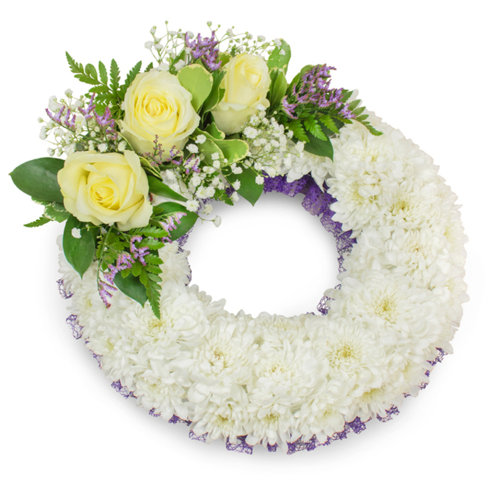 A meticulously arranged circular wreath is photographed against a stark white backdrop, its silhouette precise and its surface richly textured. The main volume consists of tightly grouped white spray chrysanthemums, each small bloom contributing layered, feathered petals to form a continuous, cushion-like ring. The white heads are so uniformly placed that the wreath reads as a single soft plane, illuminated by broad, even light that reveals the gentle folds and casts a soft shadow beneath, giving the arrangement a sense of weight and physical presence. Fine purple netting or ribbon is tucked discreetly beneath the chrysanthemums along the inner and outer edges, creating a faint lavender border that adds depth and a subtle colour anchor to the design.

Breaking the perfect circle at the upper-left quadrant is a tasteful cluster of accents: three pale yellow roses with velvety, spiralled petals provide a warm focal point, set among delicate sprays of white gypsophila and small lavender statice that introduce lacy texture and a restrained contrast. Multiple types of greenery - glossy broad leaves juxtaposed with lighter, fern-like fronds - frame and support the floral cluster, enhancing both the silhouette and tactile variation. The arrangement conveys a composed solemnity, appropriate for memorial services or a graveside tribute, and evokes the calm dignity often requested for ceremonies in and around Grove Park. The overall impression is one of respectful craftsmanship, balancing purity, gentle colour, and thoughtful texture.