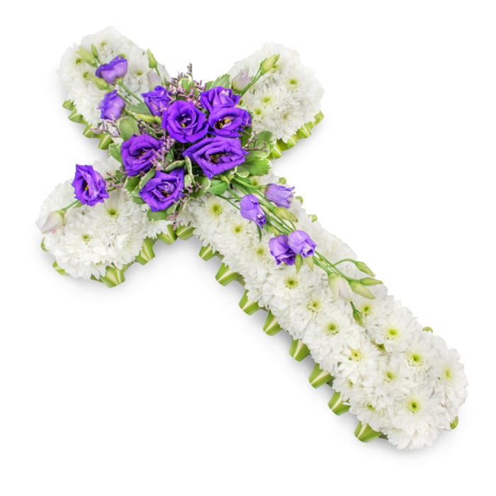 In this image, a carefully composed floral cross rests against a clean white backdrop, its simplicity and grace reminiscent of a quiet chapel near Grove Park station. The base of the cross is completely hidden beneath a densely packed blanket of white spray chrysanthemums, their small blooms pressed closely together so that the surface appears almost plush to the touch. Each chrysanthemum is in full flower, with crisp white petals fanning out around a tiny green centre, creating a gentle pattern that repeats rhythmically across the arms and length of the cross. Tracing the outer edges is a slender lime-green ribbon, meticulously pinned to follow the contours of the cross and provide a defined, contemporary frame that subtly lifts the design, much like a modern accent in a traditional church interior. At the central junction, where the vertical and horizontal arms meet, a lush cluster of royal purple lisianthus commands attention. These flowers, with their layered, slightly ruffled petals, range from fully open blooms revealing rich velvet depths to tightly furled buds promising to open. Slender stems of light green foliage and delicate filler flowers weave among the lisianthus, softening the outline and adding fine texture. From this core, several lisianthus stems drift diagonally down the lower-right arm of the cross, creating a gentle cascade that brings movement and visual flow, echoing the meandering path of the nearby Thames. The lighting is bright but diffused, producing a faint halo of shadow beneath the cross that lends subtle depth without distracting from the arrangement itself. This floral cross conveys a sense of calm faith, dignity, and condolence, an ideal tribute for services held in Grove Park churches, crematoria, or local cemeteries.