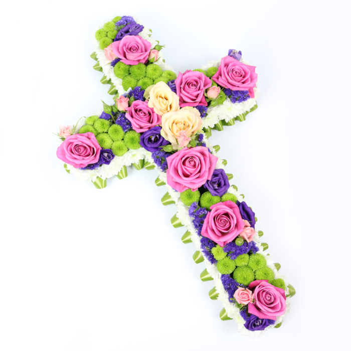 A beautifully detailed floral cross rests against a bright white backdrop, photographed from a slightly elevated angle that reveals both its precise outline and generous depth. The cross is meticulously edged with a dense border of petite white spray chrysanthemums, each bloom nestled closely to its neighbour to create a continuous, velvety band of white. Just beyond this soft fringe, a slim band of folded light green ribbon or foliage runs all the way around, adding a gentle hint of colour and structure that calls to mind the calm green spaces and tree-lined avenues of Grove Park. Within this delicate frame, the interior of the cross is a vibrant mosaic of roses, button chrysanthemums, and smaller accent flowers. Dominating the design are large roses in rich fuchsia pink and pale, creamy yellow, fully opened so their layered petals form lush spirals of colour. These roses are placed thoughtfully along the central line and across the outstretched arms, creating natural focal points. Between them, rounded lime-green button chrysanthemums punctuate the arrangement, their tight, pom-pom heads giving a lively contrast in both tone and texture. Woven among these larger blooms are clusters of small, deep purple or violet flowers-likely lisianthus or statice-that add depth, shadow, and a subtle note of dignity, well suited to a sympathy tribute. The cross is angled gently from upper left to lower right, enhancing its three-dimensional feel, as though it could be lifted straight from the image and carried to a nearby Grove Park church or cemetery. Bathed in even, bright light, every detail is crisp and visible. This floral cross expresses faith, remembrance, and an uplifting celebration of life, ideal for honouring a loved one with grace.