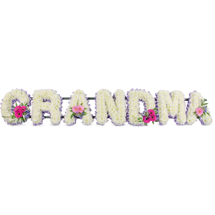 This floral tribute shows the word GRANDMA set out in a straight line of strong, block-style letters, all built on firm frames and then completely hidden under a dense layer of white chrysanthemums. Each small chrysanthemum head is positioned so tightly that their petals touch, forming a solid, padded field of white across every character, with soft curves and edges that soften the otherwise bold shapes. Surrounding each letter, a lilac ribbon has been arranged along the outer contour, gently frilled to add a decorative edge that is both tidy and delicate, giving the impression of a carefully stitched border on a sentimental keepsake. On the first G, one of the middle As, and the last A, additional floral clusters introduce a quiet sense of celebration. These clusters contain pink gerbera daisies, their round centres and radiating petals creating clear focal points, and pink roses in various stages of opening, accompanied by leafy green foliage that adds contrast and a feeling of freshness. The colour harmony is soft and understated, with pastel lilac and pink playing against the crisp white background. The lighting appears calm and evenly spread, revealing the textures of petals, ribbon, and leaves without distraction. It is easy to imagine this arrangement prepared by an experienced Grove Park florist and delivered with care to a funeral in the local area, perhaps after being designed in a studio not far from Grove Park station or the green spaces around Chiswick House. The tribute radiates affection and respect, serving as a floral embodiment of a grandmother's kindness, patience, and lasting influence on her family.