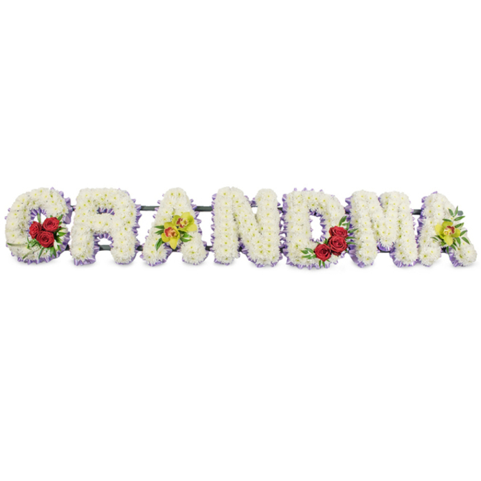 In this scene, the word GRANDMA stretches across the frame in a line of large, carefully shaped floral letters, presented against a clear white background that emphasises the simplicity and elegance of the tribute. Each capital letter is made from a dense bed of small white chrysanthemums, their petals layered tightly together until no stems or gaps are visible, creating a plush, almost cushion-like surface that suggests care and tenderness. Beneath the white blooms at the base of each letter runs a delicate edging of pale lavender or soft purple flowers, offering a subtle contrast and lending a sense of depth, like a faint shadow of colour underneath the clean white. The first letter, G, is enriched by a cluster of three deep red roses placed on its lower left curve, their velvety petals full and open, surrounded by dark green leaves that curl softly around them. On the first A, two yellow-green orchids are positioned along the crossbar, their chartreuse petals and darker centres creating a refined, slightly exotic highlight. The middle letters R, N, and D remain unadorned beyond their white petals and lavender trim, steady and serene. The M displays another trio of red roses nestled within its lower right dip, a thoughtful echo of the G, and the final A copies the first with two more chartreuse orchids resting within its structure. A slim dark green or black metal frame links the letters from behind, offering strength while staying unobtrusive. This floral word tribute feels deeply personal, the kind of arrangement a family in Grove Park might order to accompany a hearse or rest beside a coffin, letting the calm whites, soft purples, and warm reds express love, gratitude, and farewell to a much-loved grandmother.