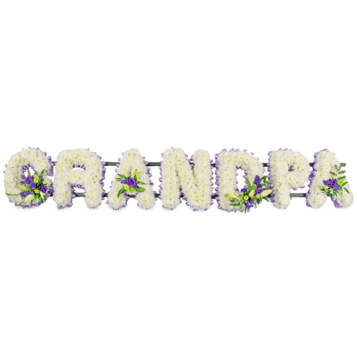 Shown here is a personalised funeral tribute spelling out GRANDPA in clear, upright capital letters, positioned horizontally as though ready to be placed beside a coffin or at the front of a chapel in Grove Park. Each letter is meticulously formed from an abundance of white chrysanthemums or carnation-style flowers, packed so closely that the surface resembles a plush floral cushion. The texture is rich and inviting, with the many petals overlapping to create soft highlights and gentle shadows that contour the curves and angles of each character. Against this pure white canvas, accents of rich purple and soft lavender blooms appear at carefully chosen points, paired with sprigs of fresh green foliage that introduce a soothing, natural contrast. These accent sprays rest in the hollow of the G, nestle into the triangular centres of the As, and decorate the D, P, and final A, adding rhythm and movement as the viewer's eye travels across the tribute. A narrow dark green bar beneath the flowers links the letters together, largely hidden from view yet vital for structural support. The result is an arrangement that feels both strong and tender, the kind of bespoke piece Grove Park Florist might prepare for a family gathering to say goodbye. It would look at home in a local church service, a crematorium chapel, or a quiet memorial space overlooking the trees, offering a visual expression of respect, affection, and the enduring role a grandfather plays within the family's story.