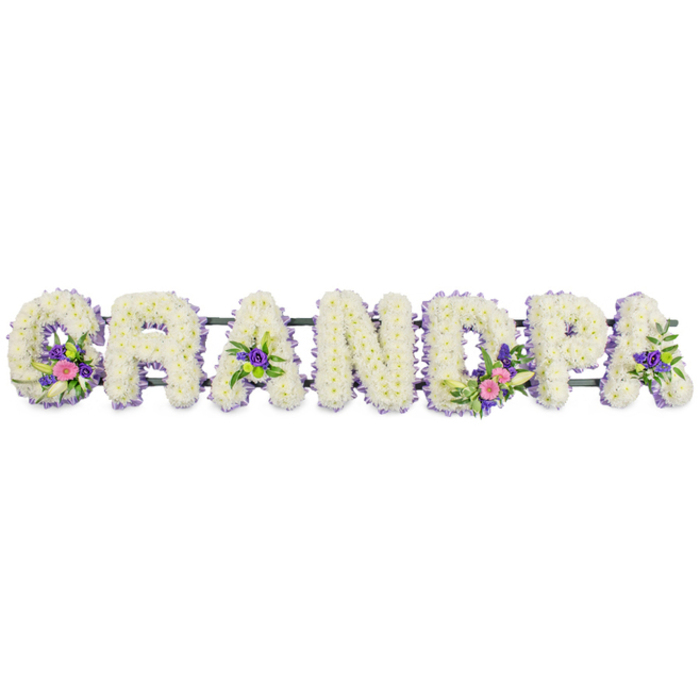 This photograph shows a bespoke floral tribute that spells out GRANDPA, the kind of piece many families in Grove Park choose when they want a deeply personal funeral arrangement for a much-loved grandfather. The letters are large, upright, and clearly legible, each one meticulously covered in tightly clustered white chrysanthemums, creating a soft, pillowy texture across the front of the tribute. The white flowers appear freshly opened, their petals clean and bright, symbolising remembrance and peace. To define the outline of each capital, the florist has added a delicate edging of tiny lilac and purple blooms, possibly statice or heather, running seamlessly around every curve and corner. This border frames the letters beautifully, providing a cool, pastel contrast that helps the word stand out in a chapel or crematorium setting near Grove Park. Several letters are also highlighted with graceful side sprays. On the 'G', an arrangement of vibrant pink and fuchsia blossoms, a single white lily-style bloom, and fresh green foliage is tied diagonally across the white base, adding a touch of colour and movement. The first 'A' and the final 'A' each carry smaller accent clusters, while the 'D' features a more substantial spray that mirrors the colours of the 'G', bringing balance to the overall design. The letters rest on a slim, dark green bar, which discreetly supports them in a straight line, ready to be placed beside a coffin, at the front of a service, or near the entrance of a church off Devonshire Road or Burlington Lane. Photographed against a plain white background with calm, even lighting, the tribute's textures and colours are clearly visible. The mood conveyed is one of heartfelt respect, warmth, and gentle farewell, expressed through flowers carefully arranged to say Grandpa with grace and tenderness.