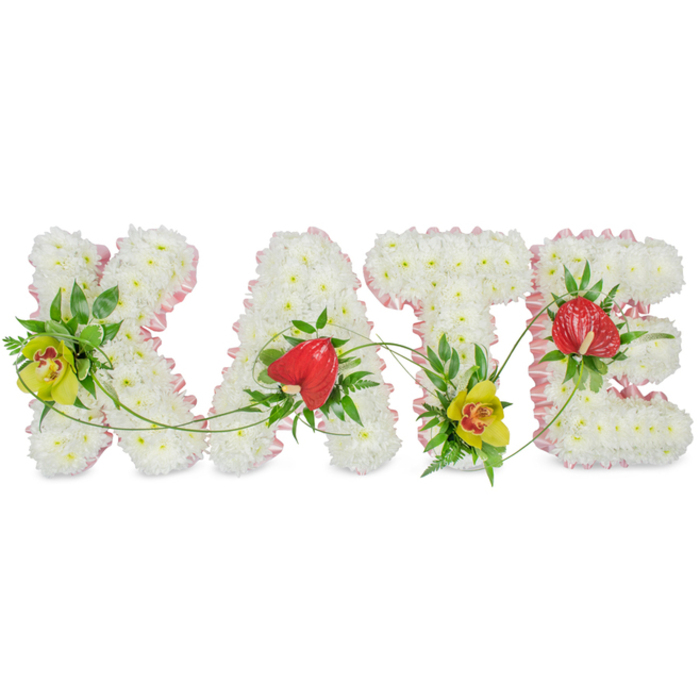 This image shows a bespoke floral arrangement spelling out the name KATE in bold, decorative letters, set against a stark white background to emphasise the clean lines and delicate detailing. Every letter is entirely covered with small, snow-white chrysanthemums, each bloom pressed closely against the next to form a dense, pillow-like surface that suggests softness and tranquillity. Along the edges, a pale pink satin ribbon is neatly pleated, outlining every angle and curve with a gentle blush of colour, softening the strong typography into something tender and personal. The left side of the K is adorned with a cluster of yellow-green cymbidium orchids, their red-flecked throats adding warmth and depth, supported by a bed of dark green foliage. A similar grouping of orchids appears at the base of the T, creating a subtle symmetry that visually balances the tribute. The A and E are accented with glossy red anthuriums, heart-shaped and slightly raised, catching the light with their waxy finish and drawing the eye to the start and end of the name. Around these focal points, varied greenery, including broad leaves, airy fronds, and slim, curling tendrils, drapes and trails over the letters, tying them together with a sense of natural movement. The design feels both structured and free-flowing, ideally suited for a funeral service or celebration of life around Grove Park, perhaps near Chiswick or along local residential streets where families gather to remember. Created with evident care, it offers a meaningful way to say goodbye to Kate through a floral tribute that feels personal, composed, and full of quiet love.